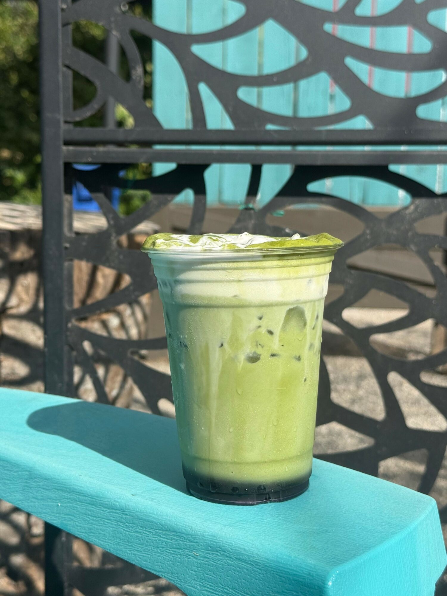 A green smoothie in a clear glass on a blue surface outdoors, with a decorative black metal fence in background.