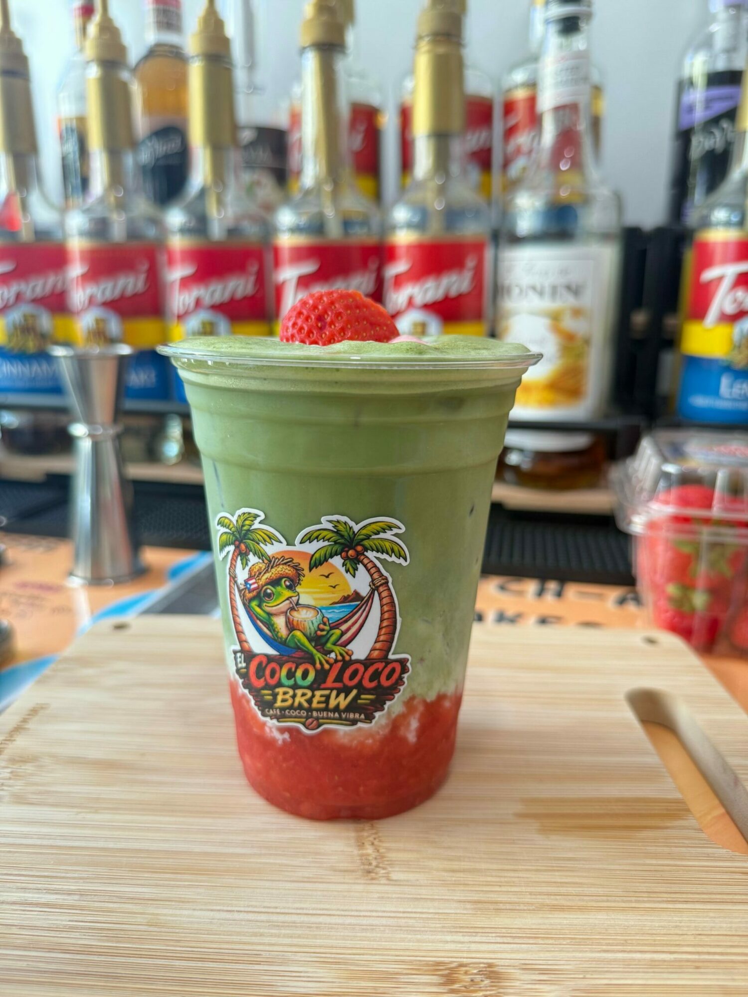 Frozen strawberry daiquiri in a green plastic cup with a strawberry garnish, on a wooden surface, with bottles in the background.