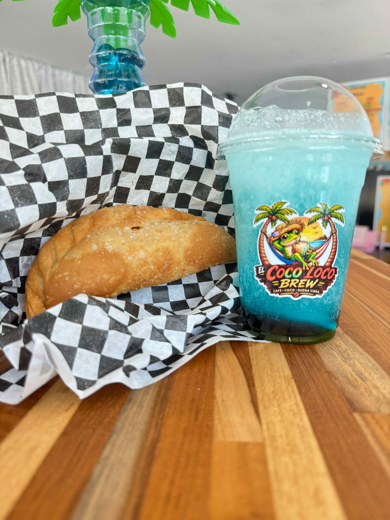 A fried empanada on black and white checkered paper, with a blue tropical-themed drink and a green plant in the background.