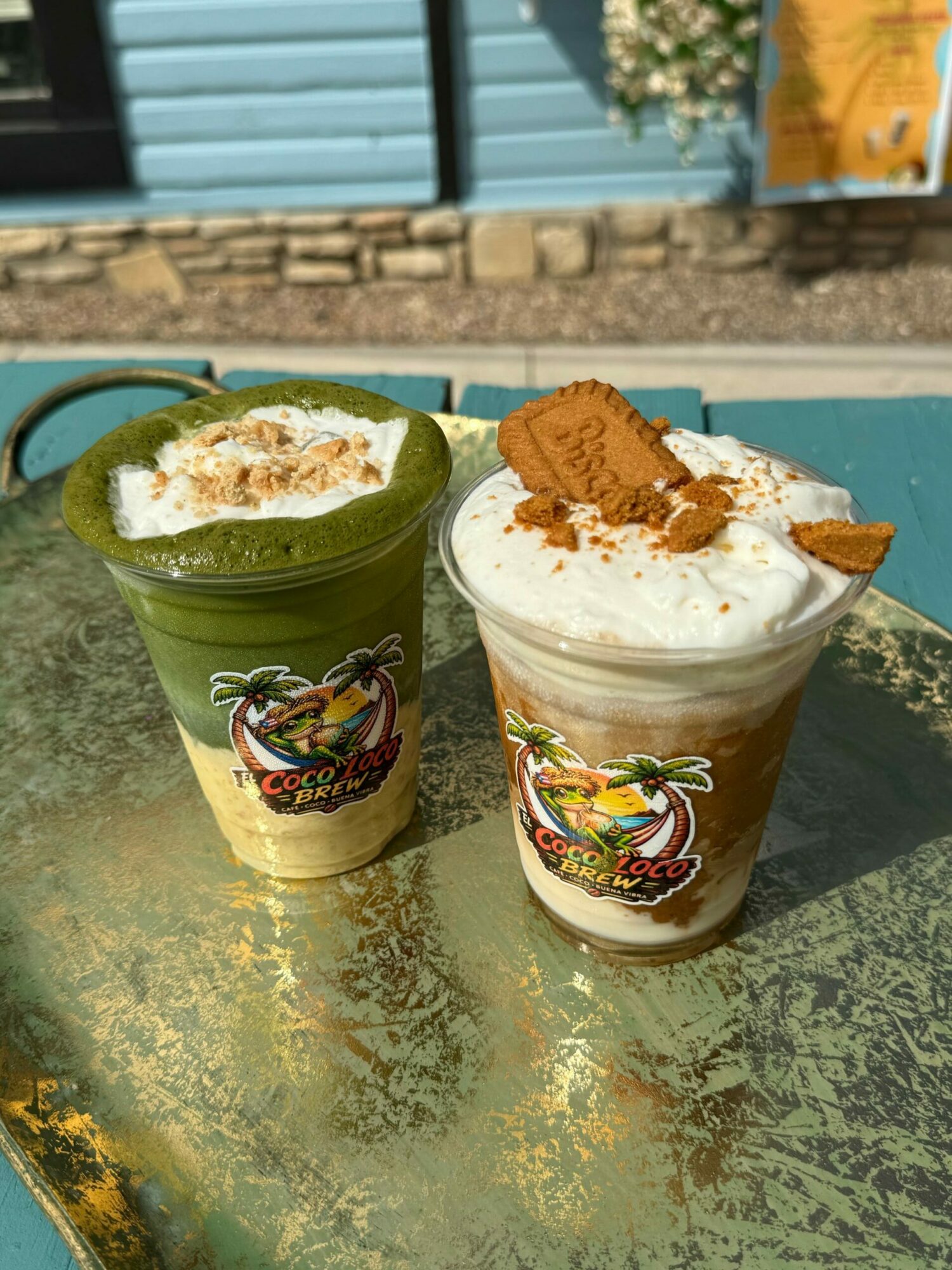 Two cups of ice cream with toppings on a green surface, one with whipped cream and cookie, the other with caramel, outdoors.