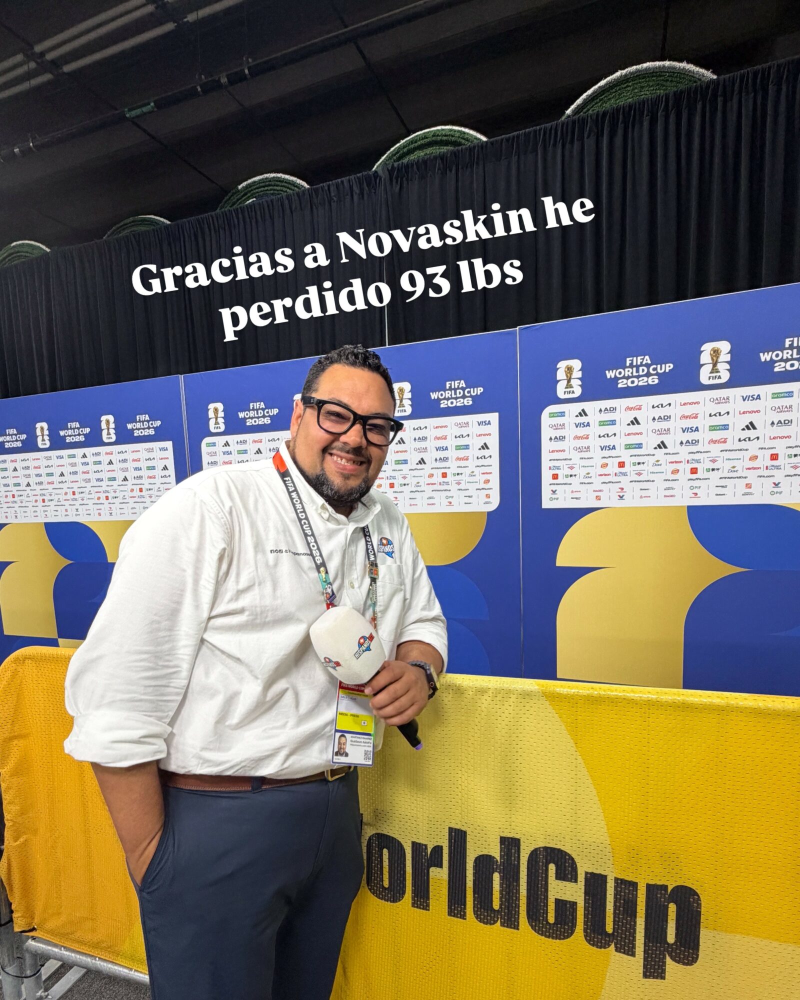 Man smiling, holding a soccer ball, standing in front of World Cup schedule boards, with a yellow barrier behind him.