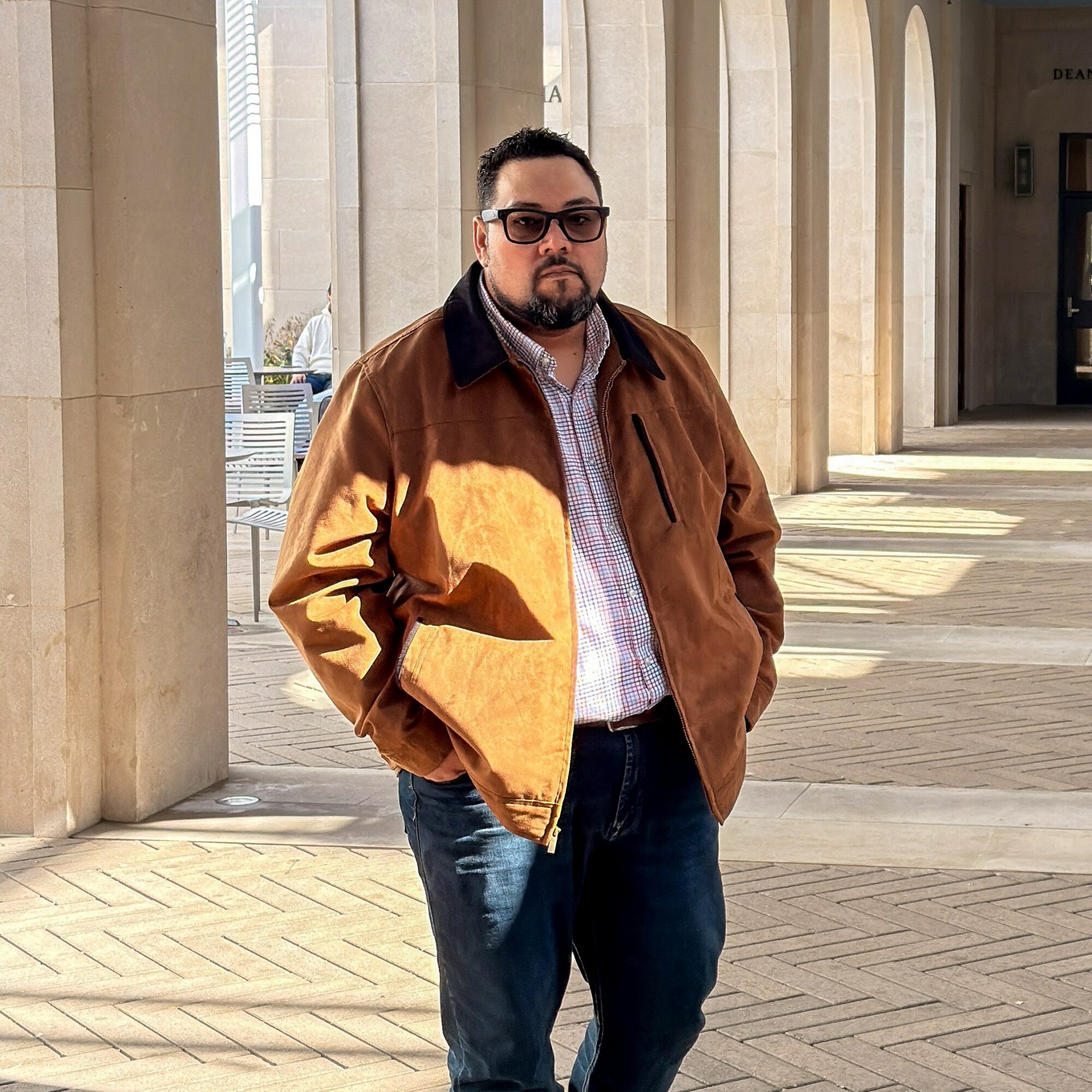 Man with sunglasses wearing a brown jacket and jeans standing outdoors near columns.