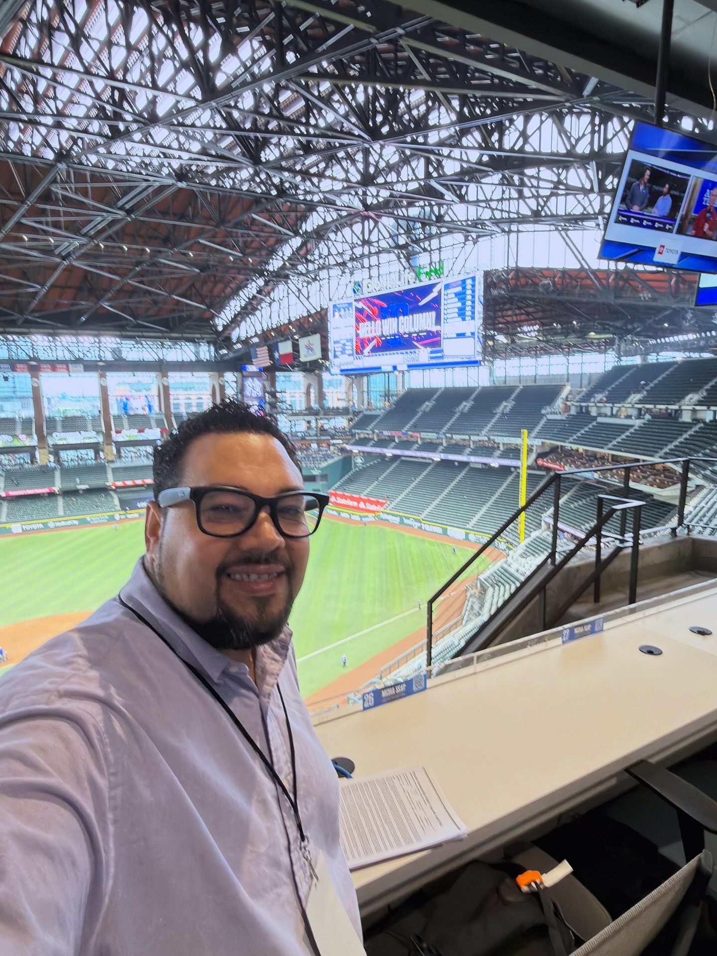Man with glasses smiling in a stadium suite overlooking a baseball field, with large screens and empty seats in the background.