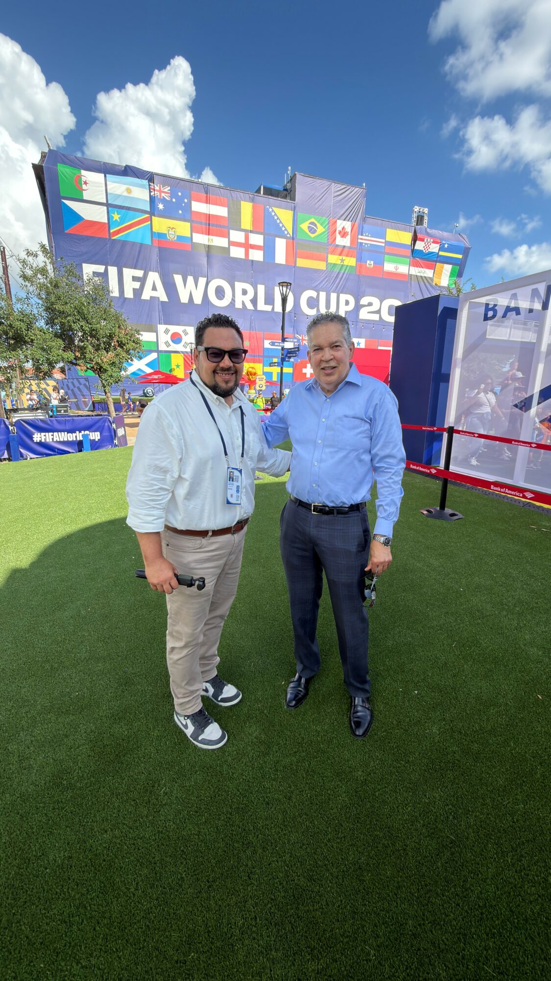 Two men standing outdoors in front of a FIFA World Cup sign, smiling, with flags and a blue sky background.