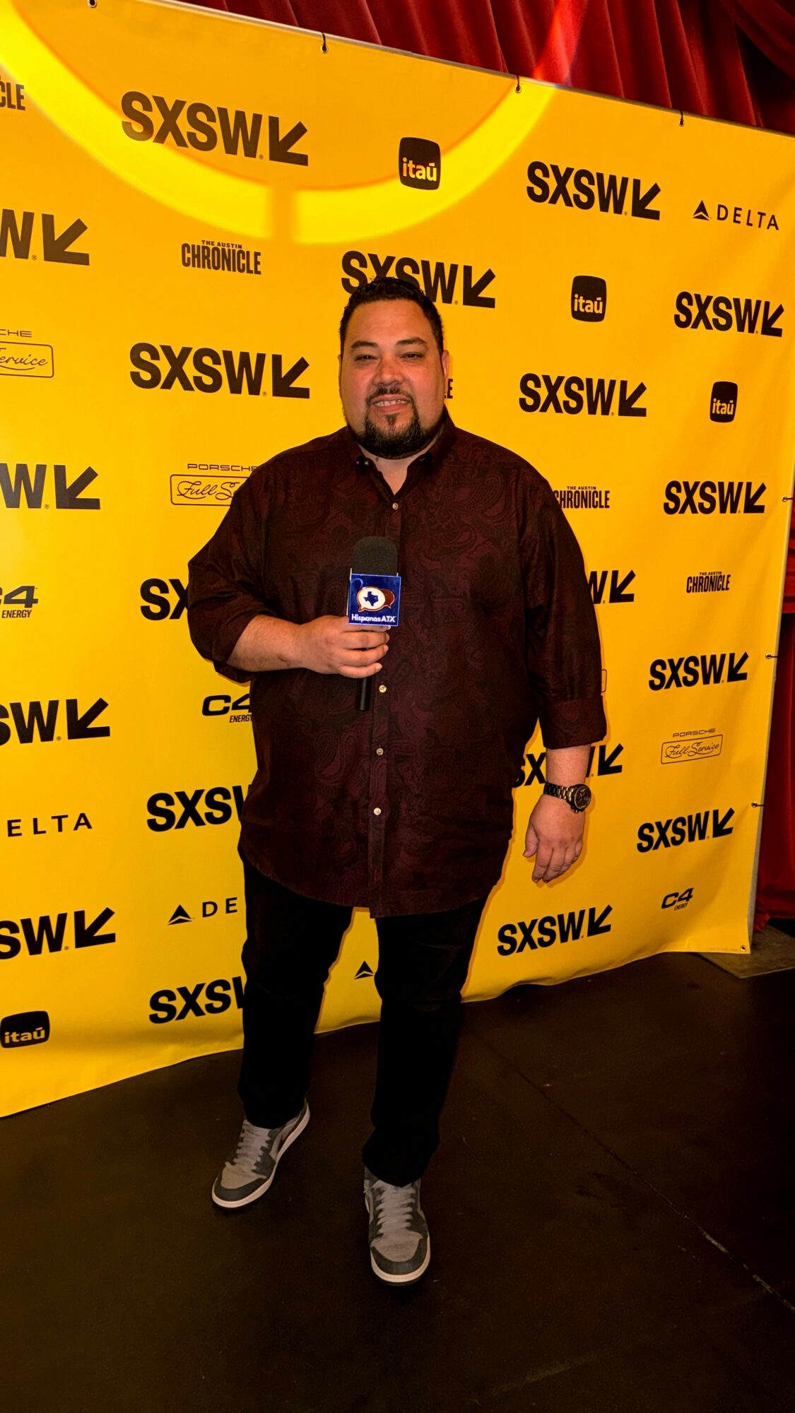 Man holding microphone standing in front of yellow SXSW backdrop with logos, smiling, wearing dark shirt and pants.