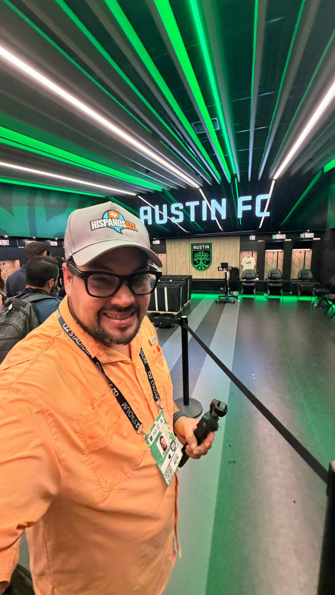 Man wearing glasses and a cap, smiling, holding a camera, standing in front of a neon-lit Austin FC sign.