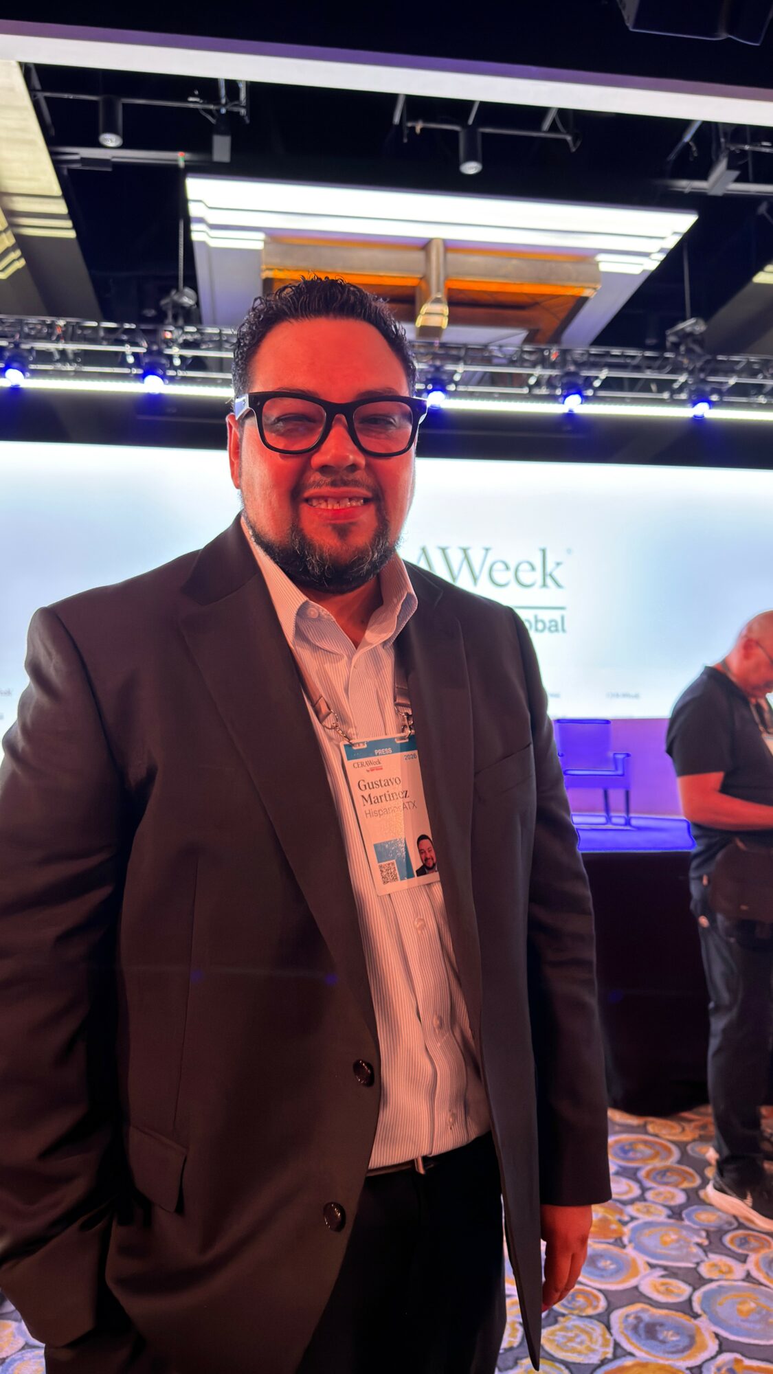 Man in suit and glasses smiling at an event with a stage and screen in the background.