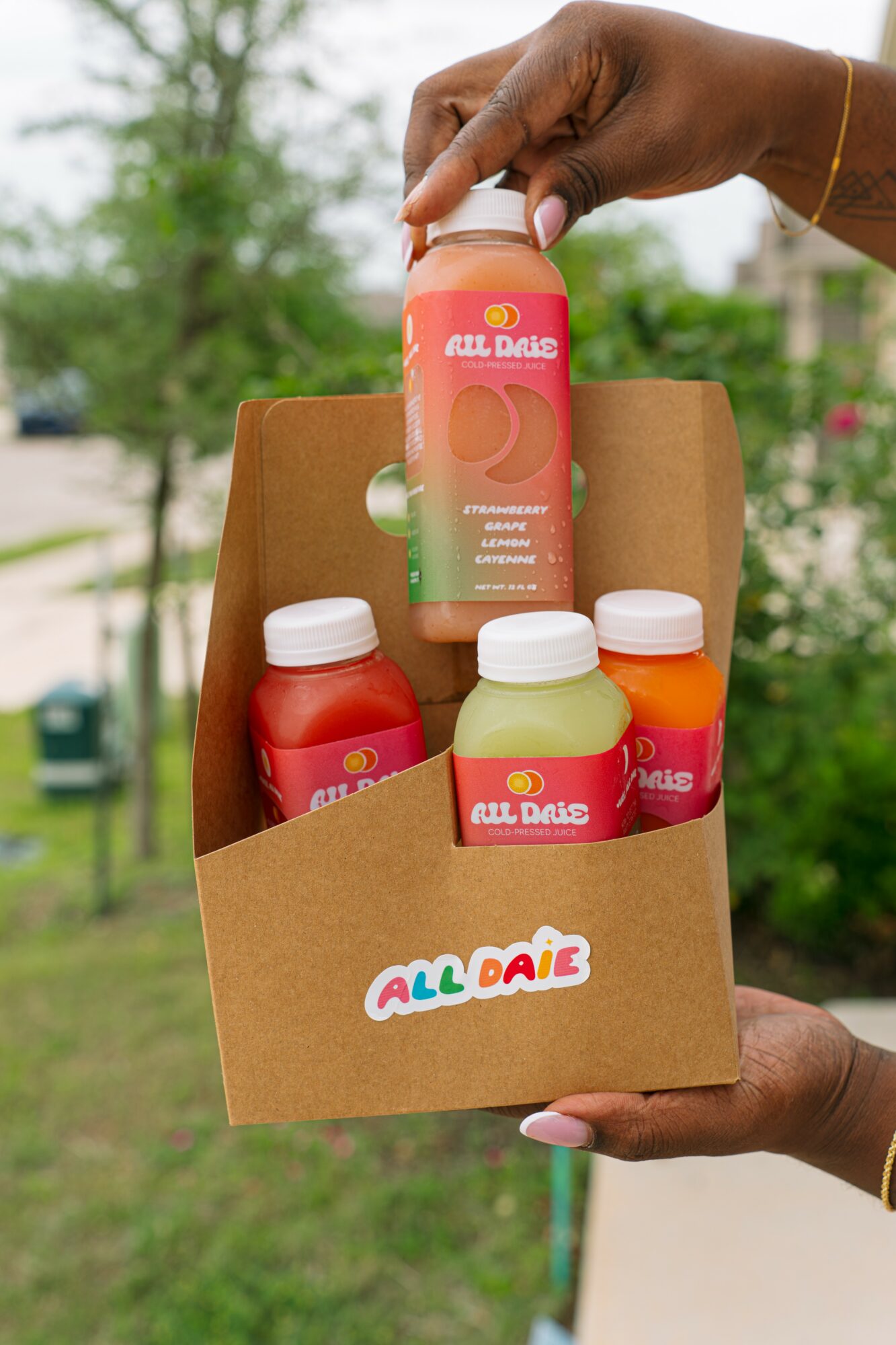 Person holding a cardboard box with colorful bottles outdoors, trees and grass in background.