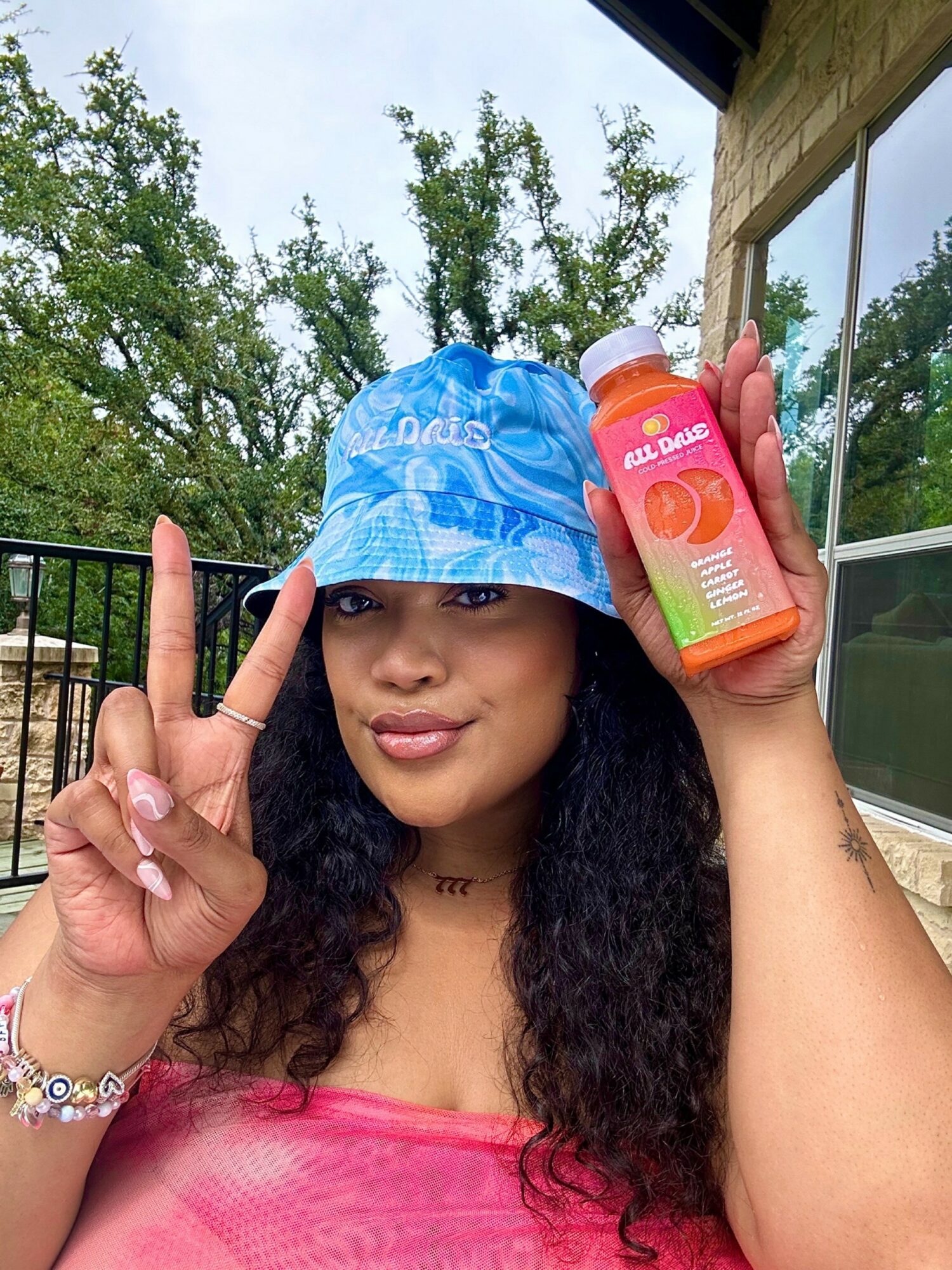 Young woman outdoors wearing a blue bucket hat, holding a peach-flavored drink, making a peace sign, smiling.