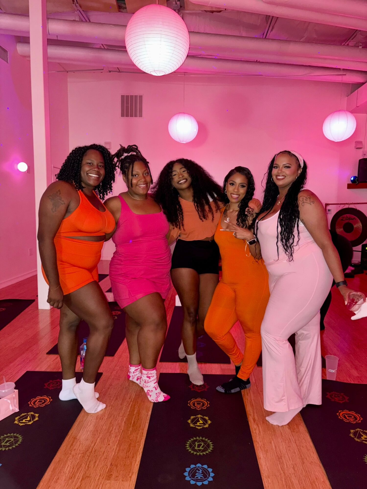 Five women pose together in a pink-lit room with hanging paper lanterns, smiling and dressed in colorful outfits.