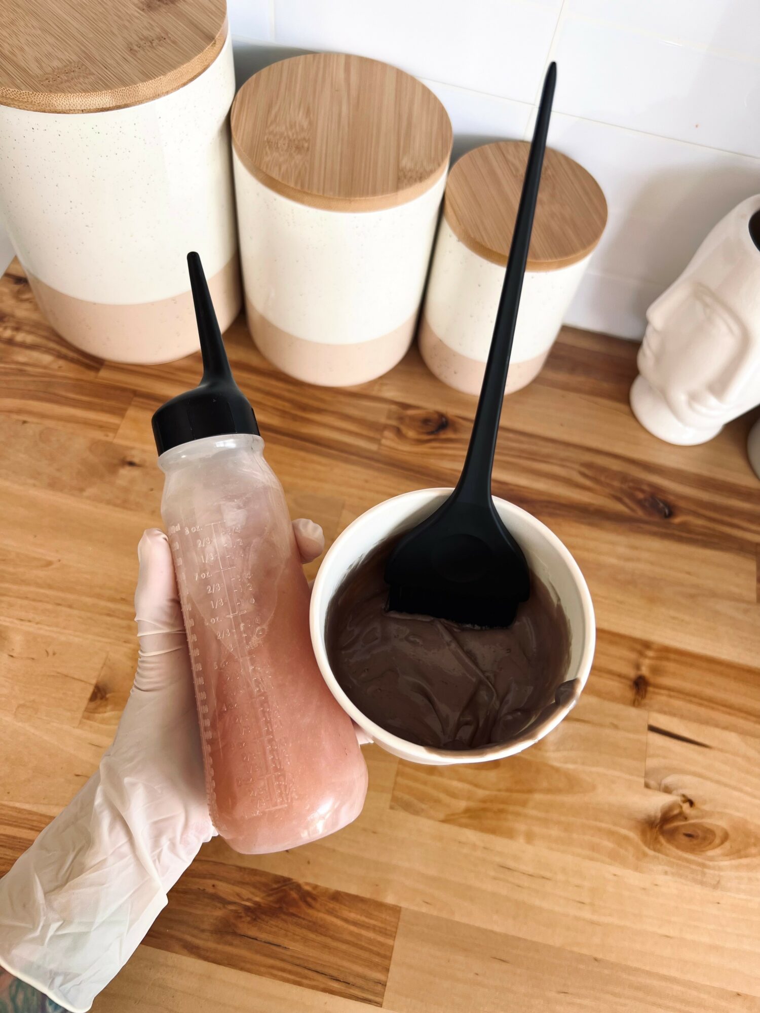 Person holding a pink smoothie in a clear bottle with a black straw, standing on a wooden floor with white and wooden containers in the background.