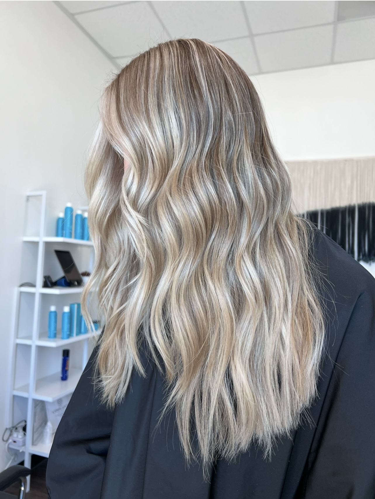 Person with long, wavy blonde hair wearing a dark jacket, standing indoors with shelves and a ceiling visible.