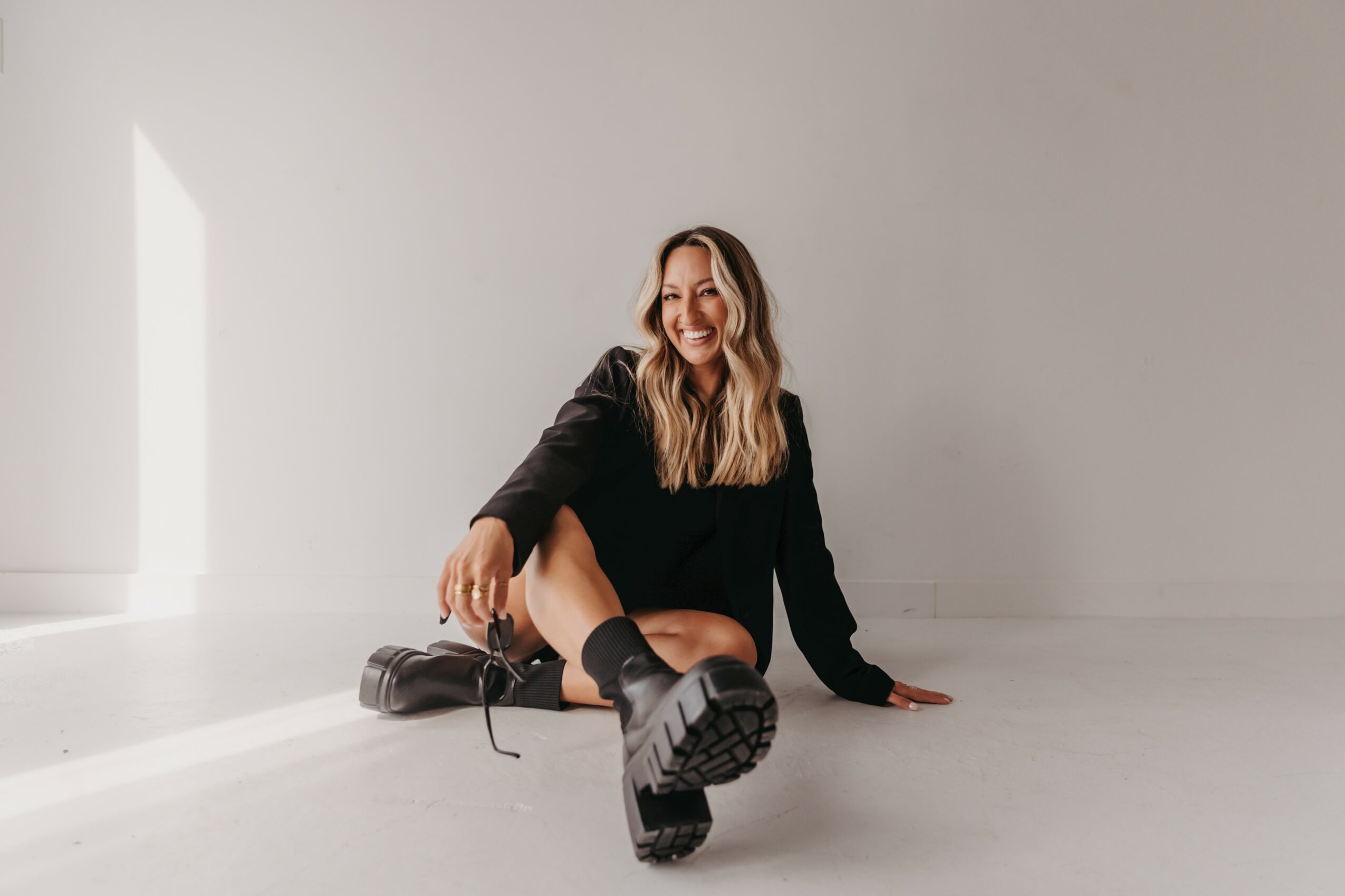 Woman sitting on the floor smiling, wearing black outfit and boots, in a bright room with white walls and floor.