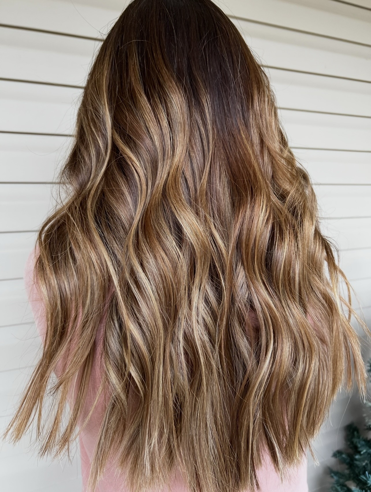 Wavy, long hair with brown and blonde highlights, viewed from the back against a white horizontal siding background.