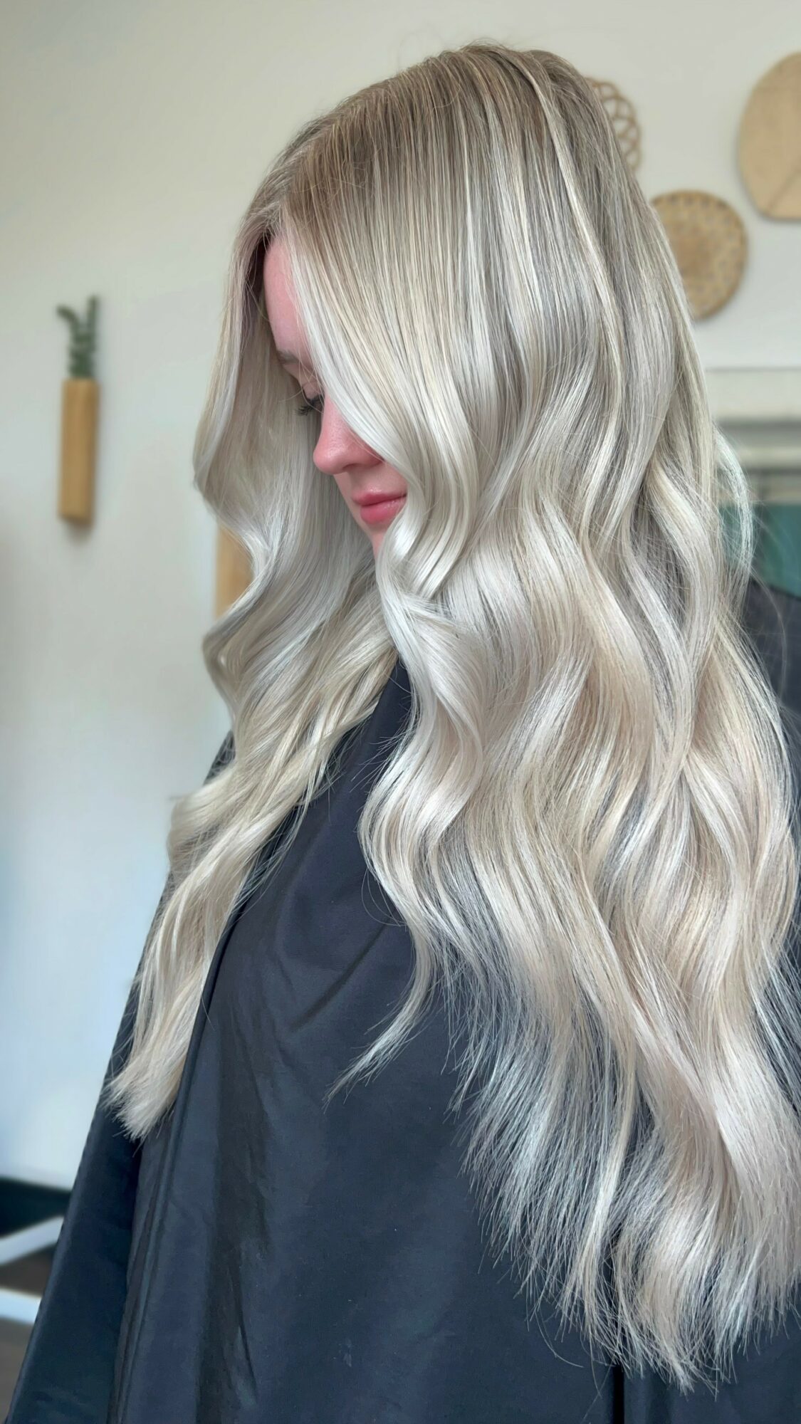 Side view of a woman with long, wavy, blonde hair, wearing a dark top, in a room with wall decor