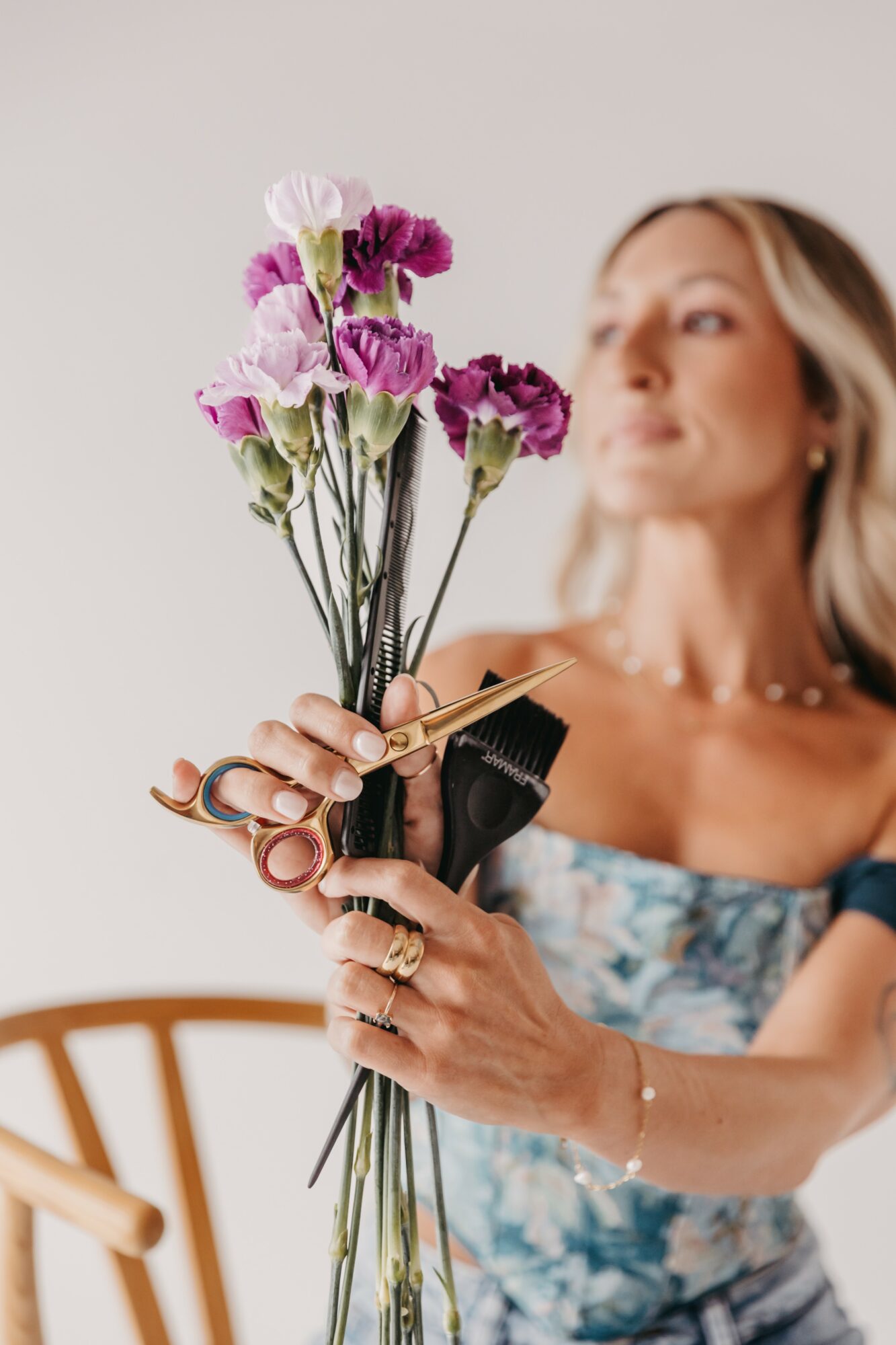 Woman holding a bouquet of pink and purple flowers, with a blurred background and a wooden chair visible.