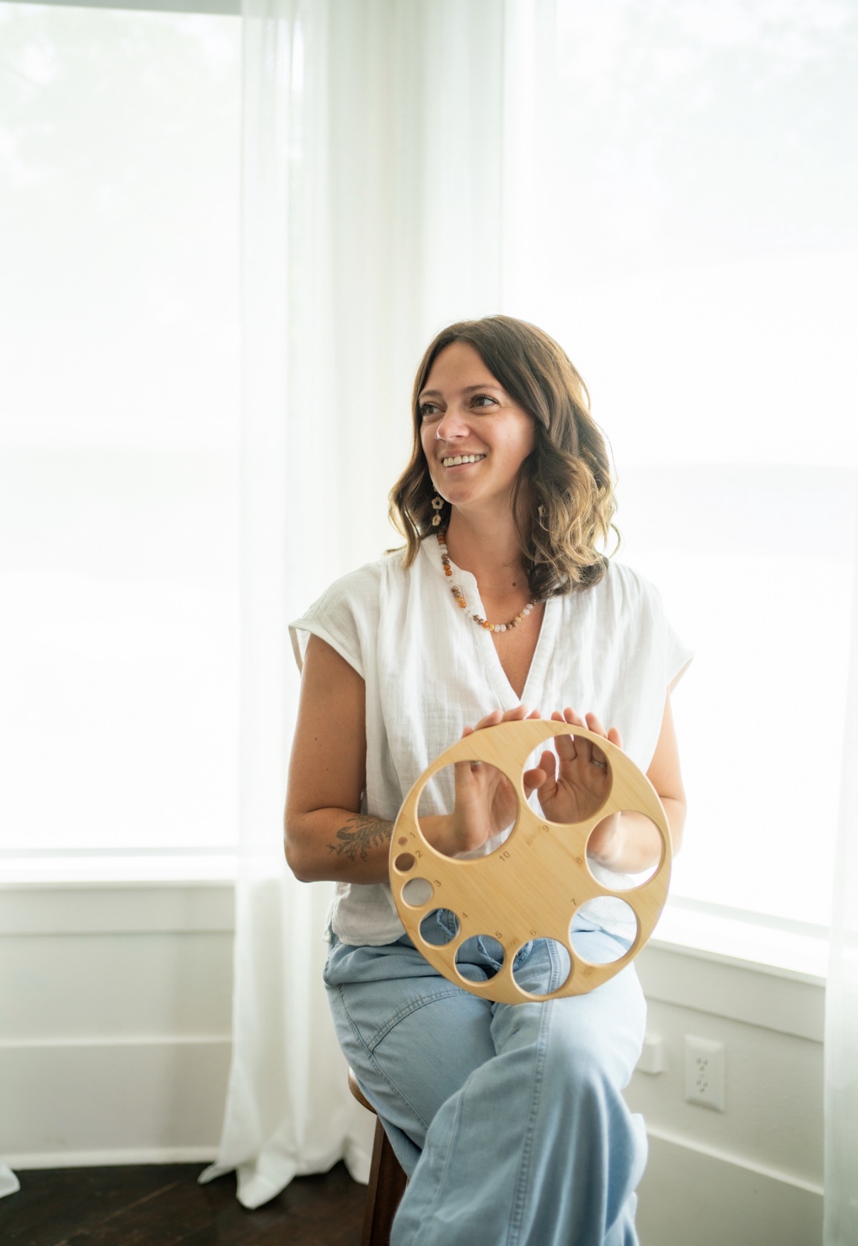 Woman sitting on a windowsill holding a circular wooden object with holes, smiling, in a bright room with white curtains.