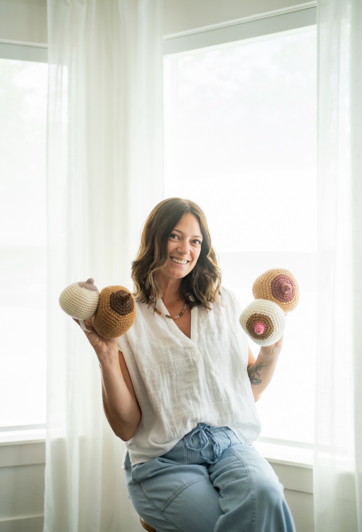Woman smiling holding colorful yarn balls in front of a bright window with curtains.
