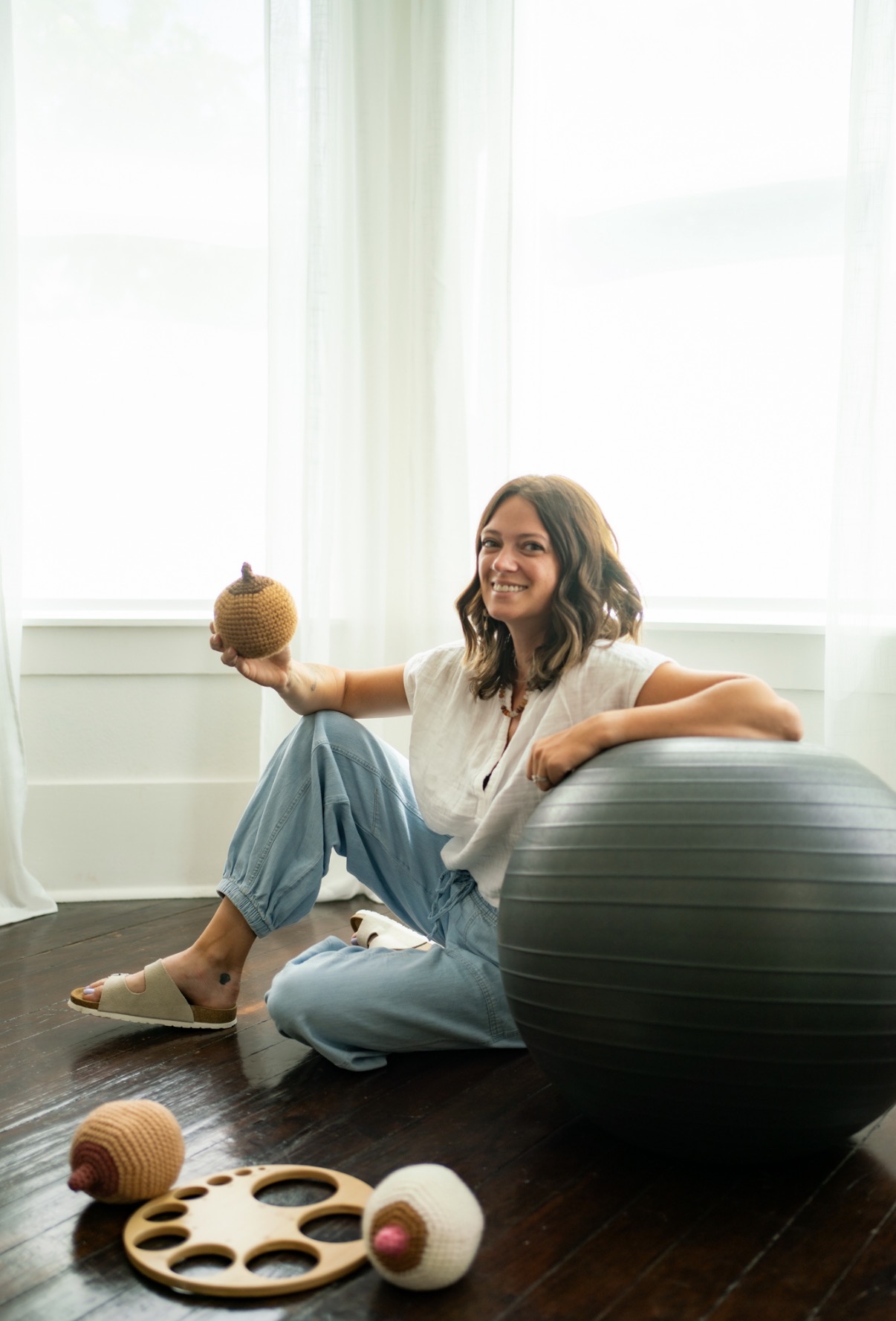 Woman sitting on floor with a large exercise ball, holding a small round object, smiling, in a bright room with large windows.