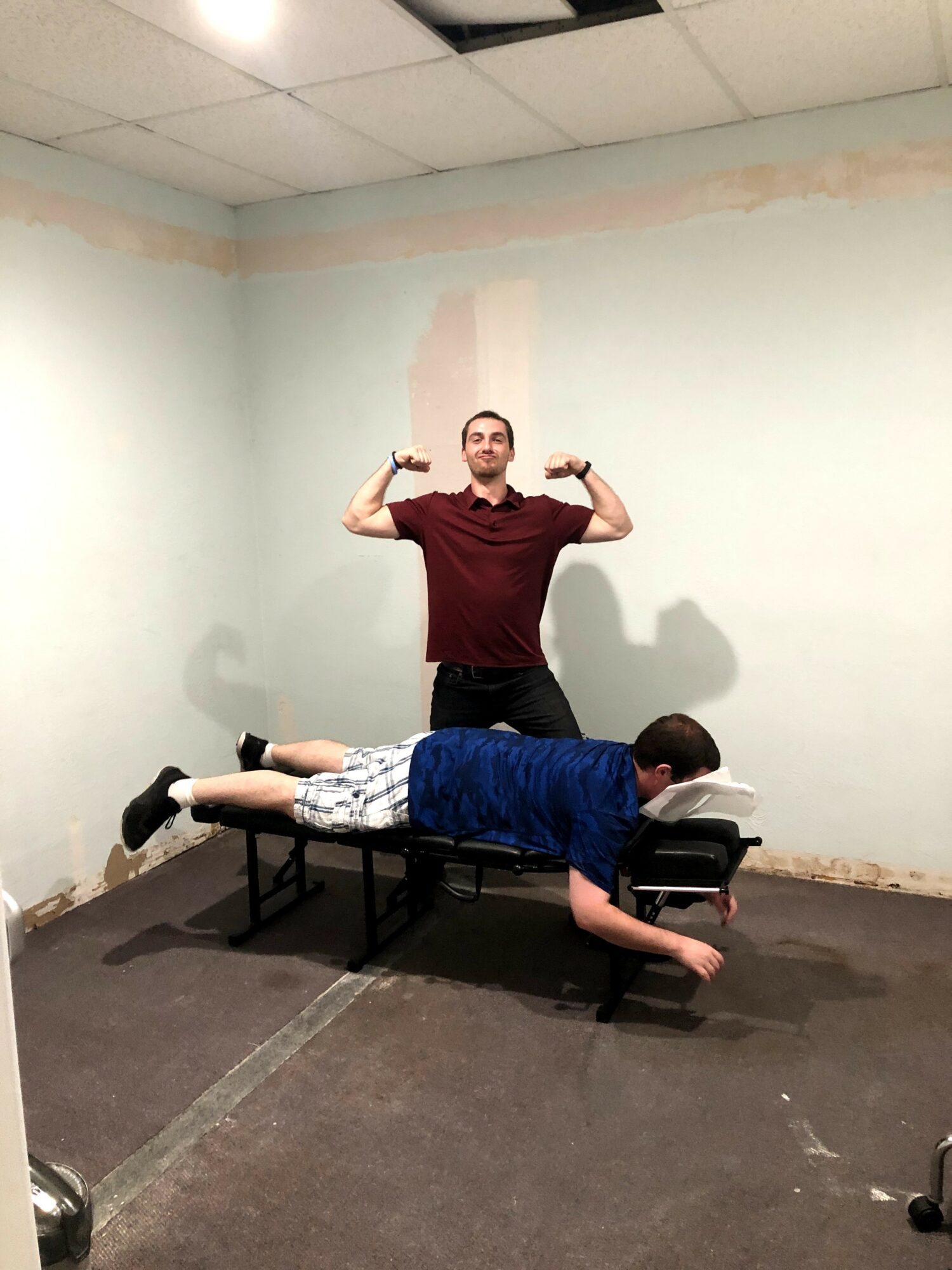 Person lying face down on a bed with a person standing behind, flexing arms, in a room with unfinished walls.