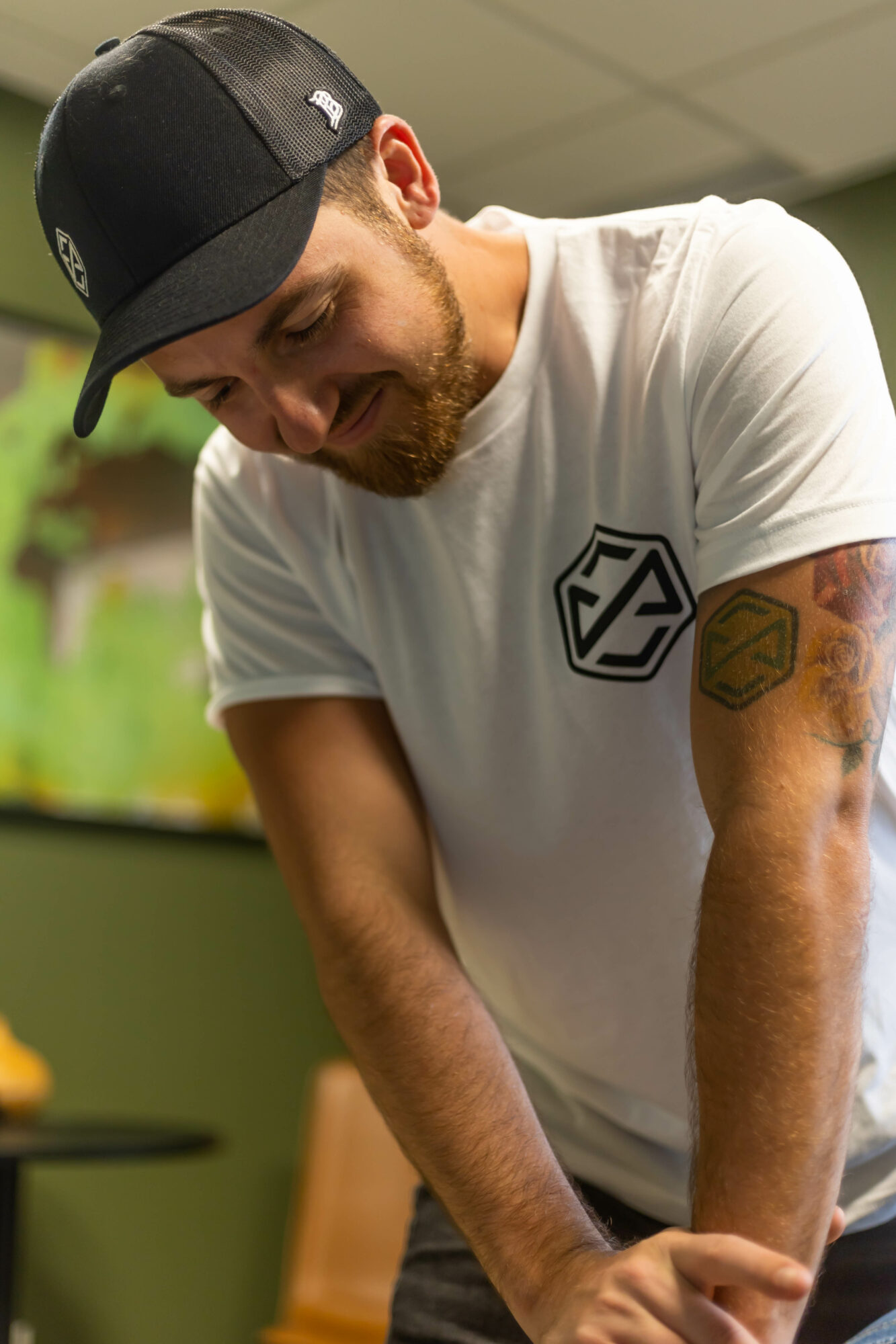 Man with a beard wearing a black cap and white t-shirt, smiling and leaning forward indoors.