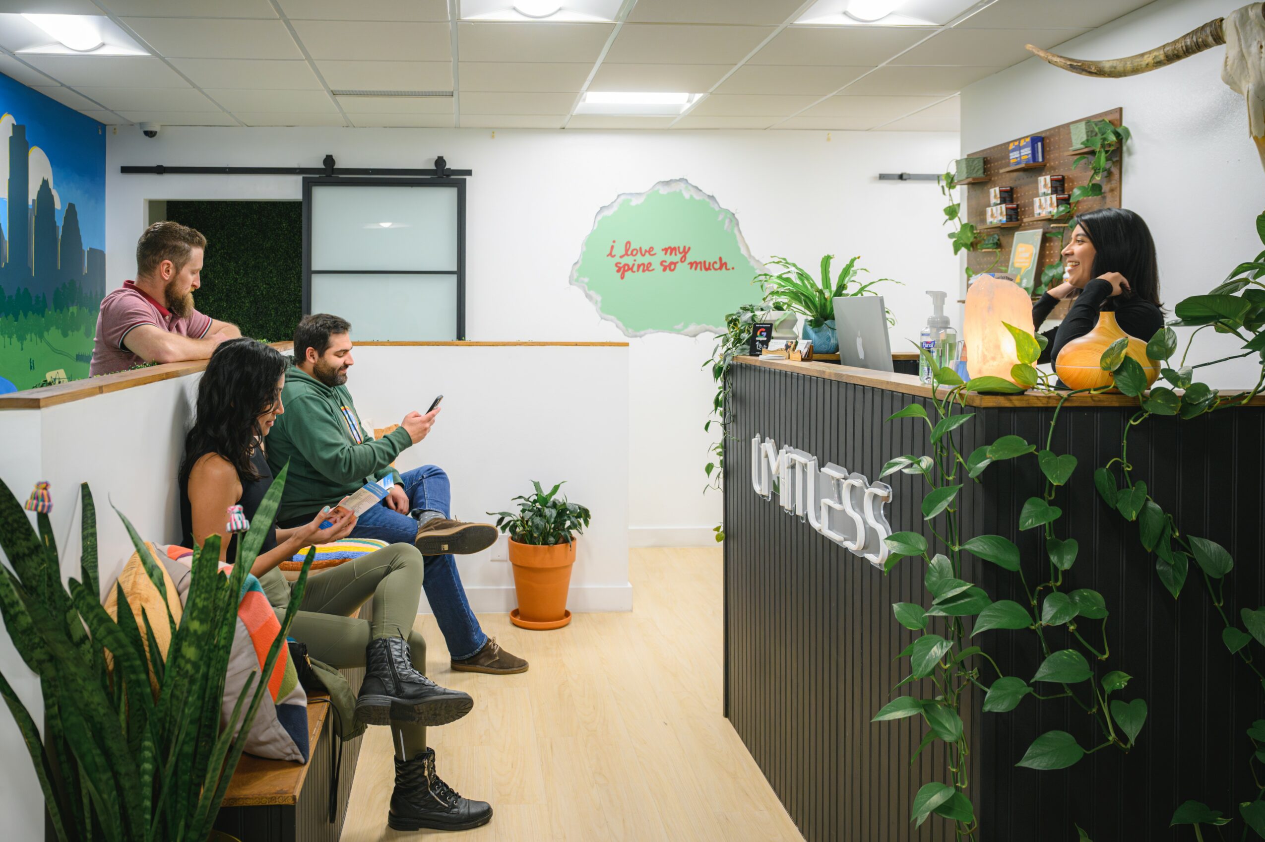 People sitting in a waiting area facing a receptionist behind a desk with plants and decorations.