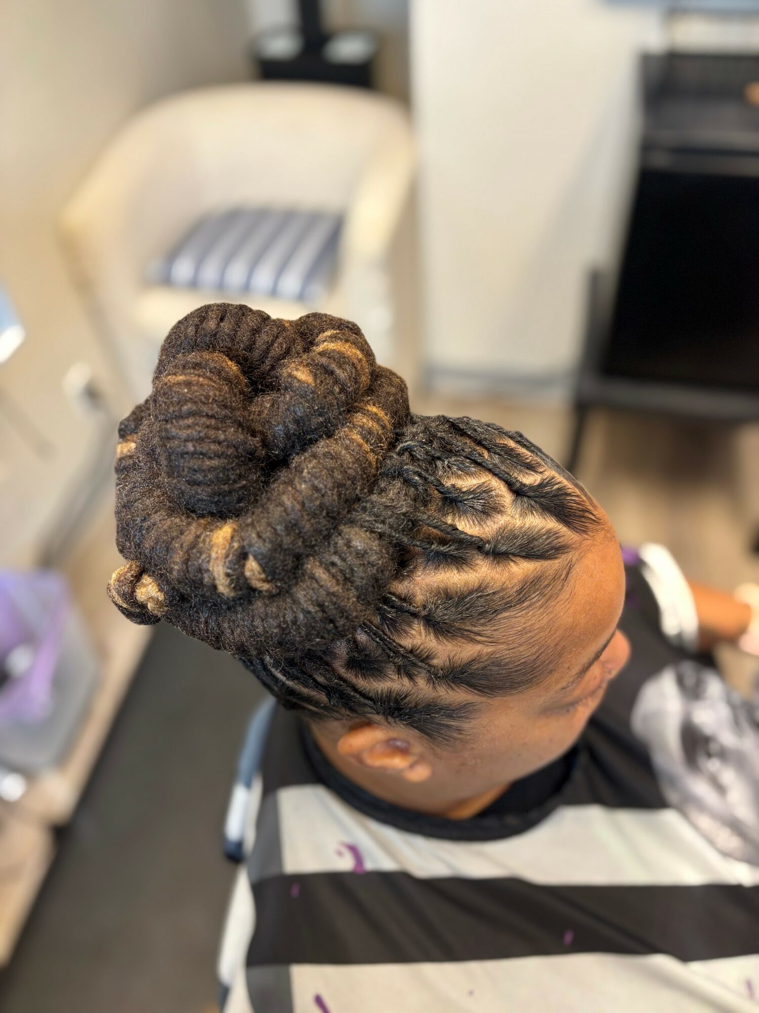 Side view of a woman with styled dreadlocks in an updo, sitting in a salon chair, background blurred.