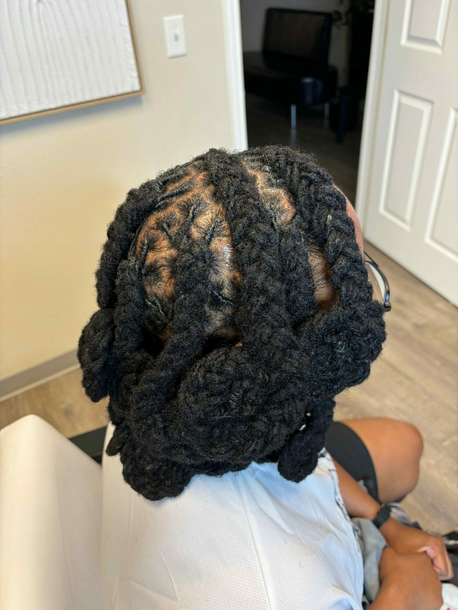 Person with dark, thick, twisted dreadlocks sitting on a chair indoors, facing away from the camera.