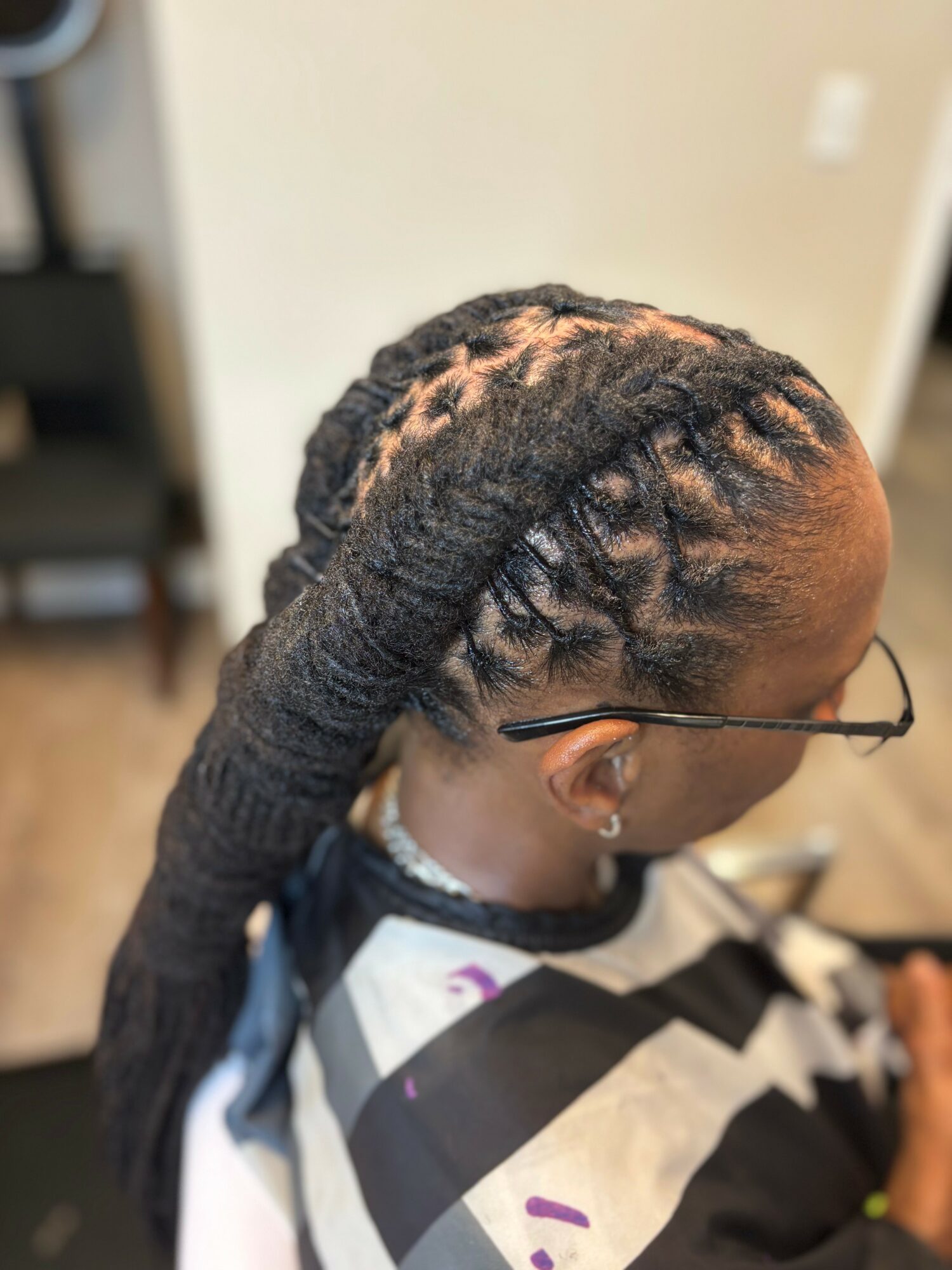 Person with long dreadlocks styled in a thick, twisted braid, wearing glasses, sitting indoors with a blurred background.