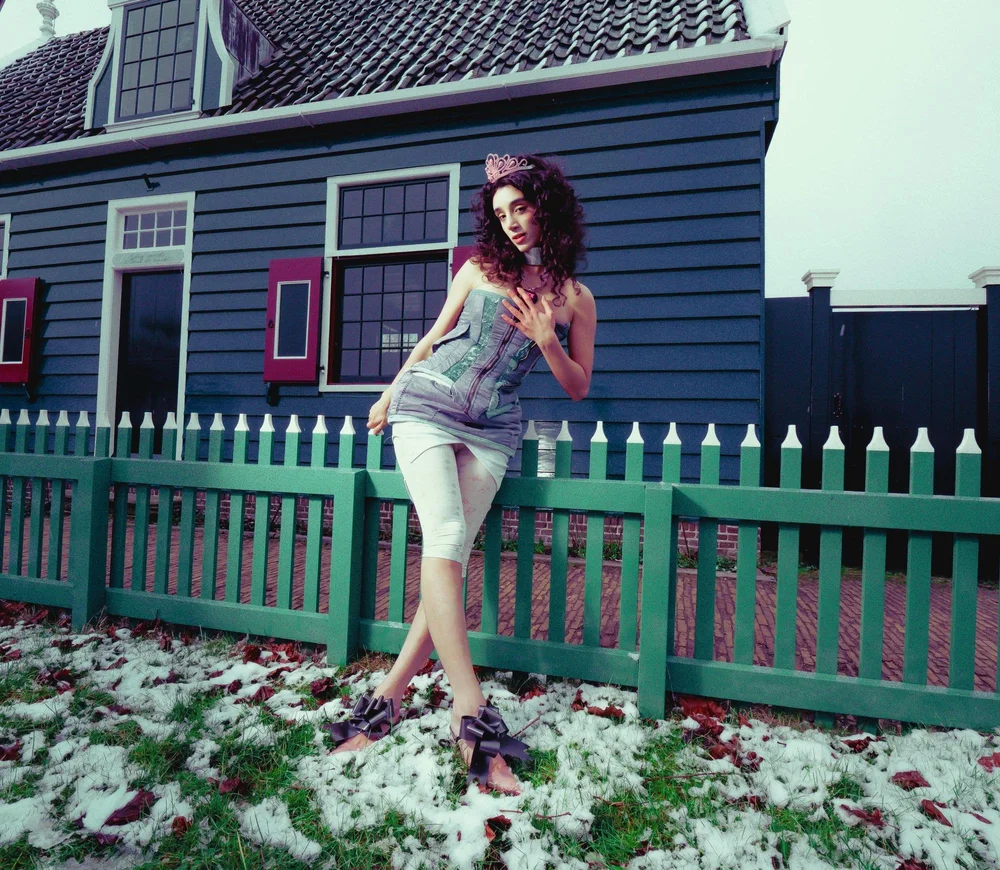 Young woman with curly hair wearing a floral headband, standing outdoors near a green fence, with a house in the background.