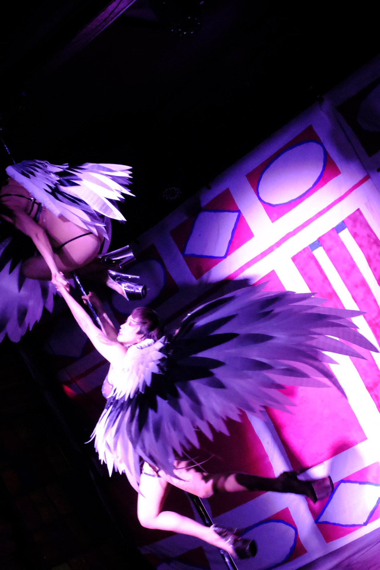 Person in costume with large feathered wings, standing on a patterned floor with purple lighting, facing away from the camera.
