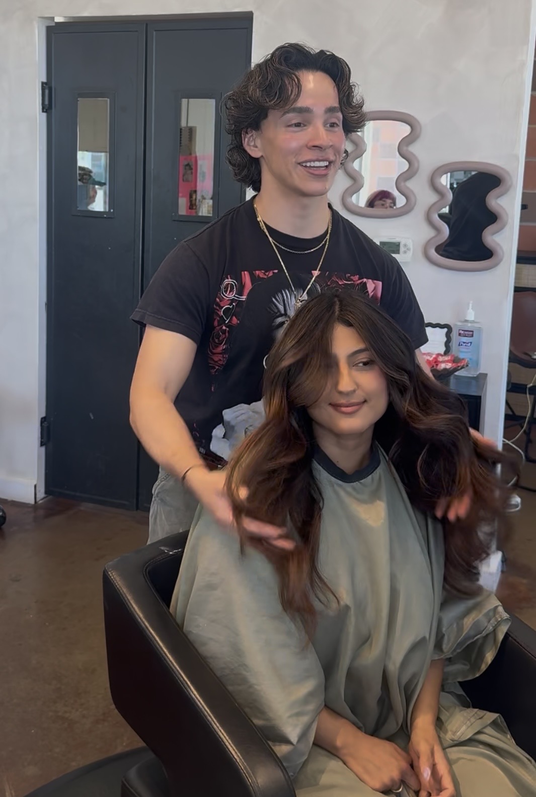 A hairstylist with curly hair stands behind a woman with long, wavy hair in a salon, smiling.