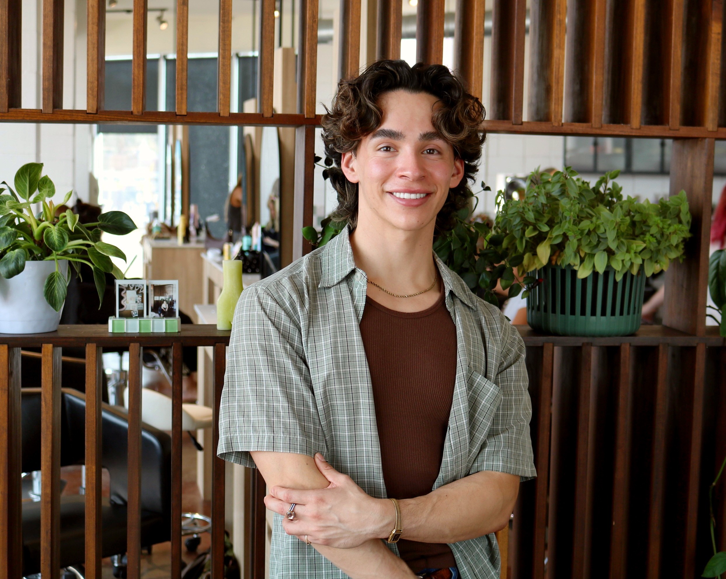 Young person with curly hair smiling, standing in front of wooden slats and potted plants.