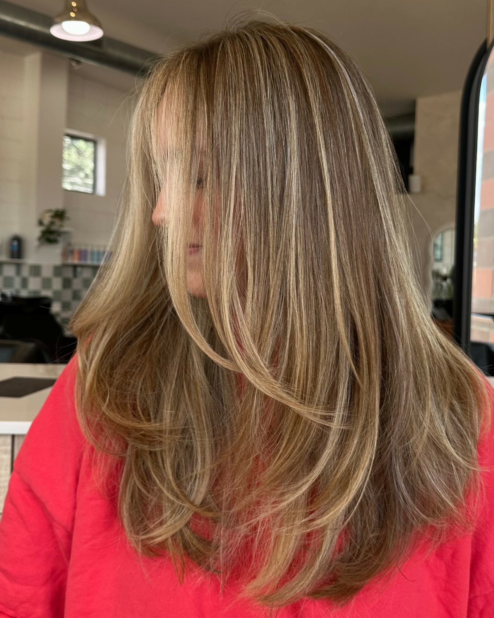 Side view of a woman with long, layered blonde hair wearing a red top, in a room with kitchen background.