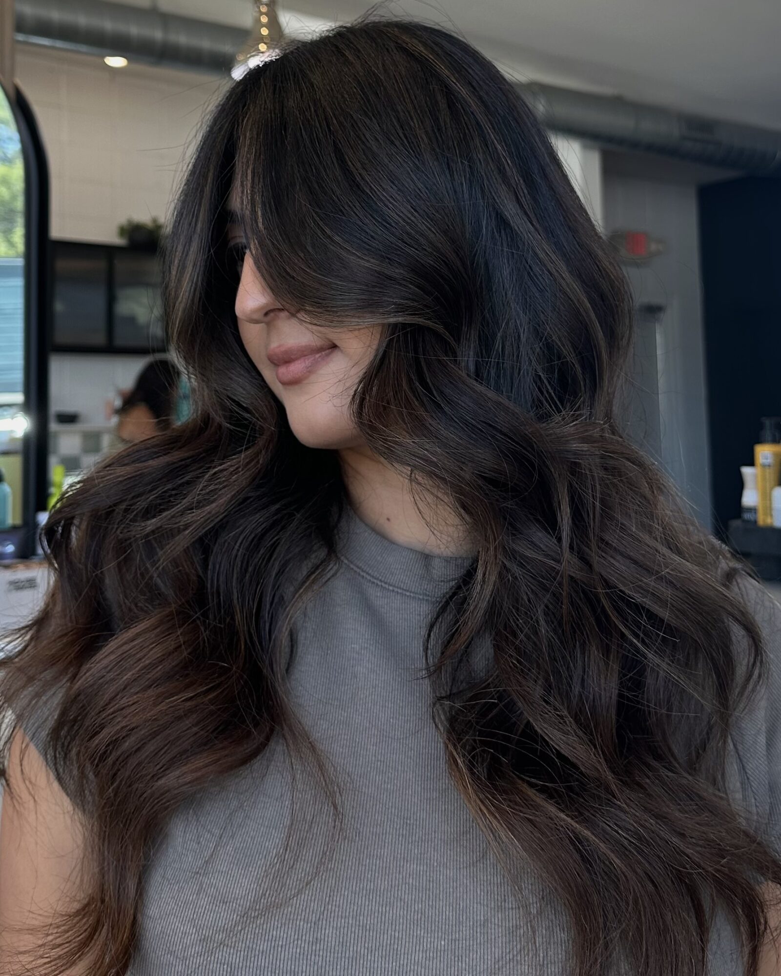 Woman with long, wavy dark hair covering part of her face, wearing a gray top, in an indoor setting.