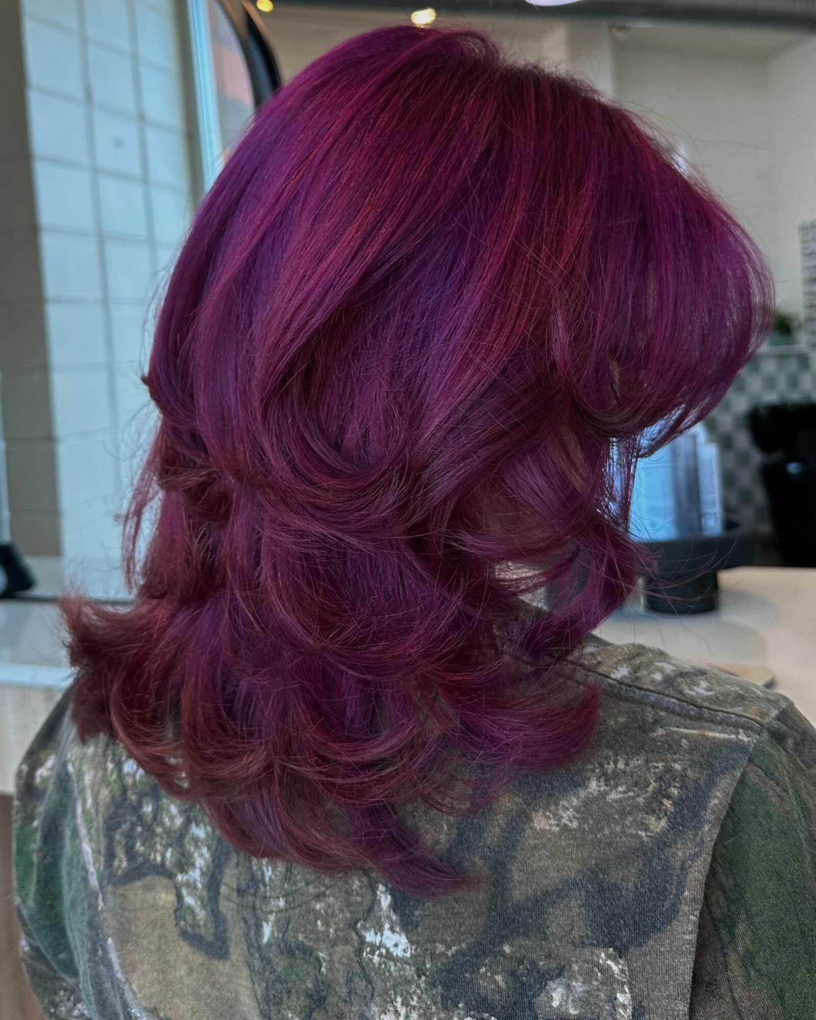Back view of a person with vibrant purple, layered, wavy hair, sitting indoors with a blurred background.