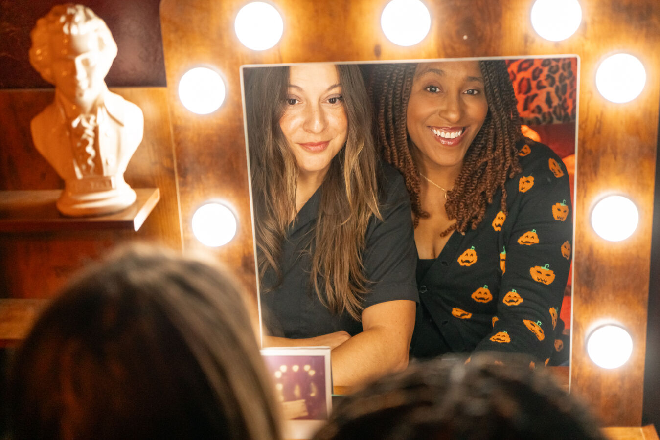 Two women smiling at a mirror with bright lights, one with long hair and the other with curly hair wearing a pumpkin-patterned shirt.