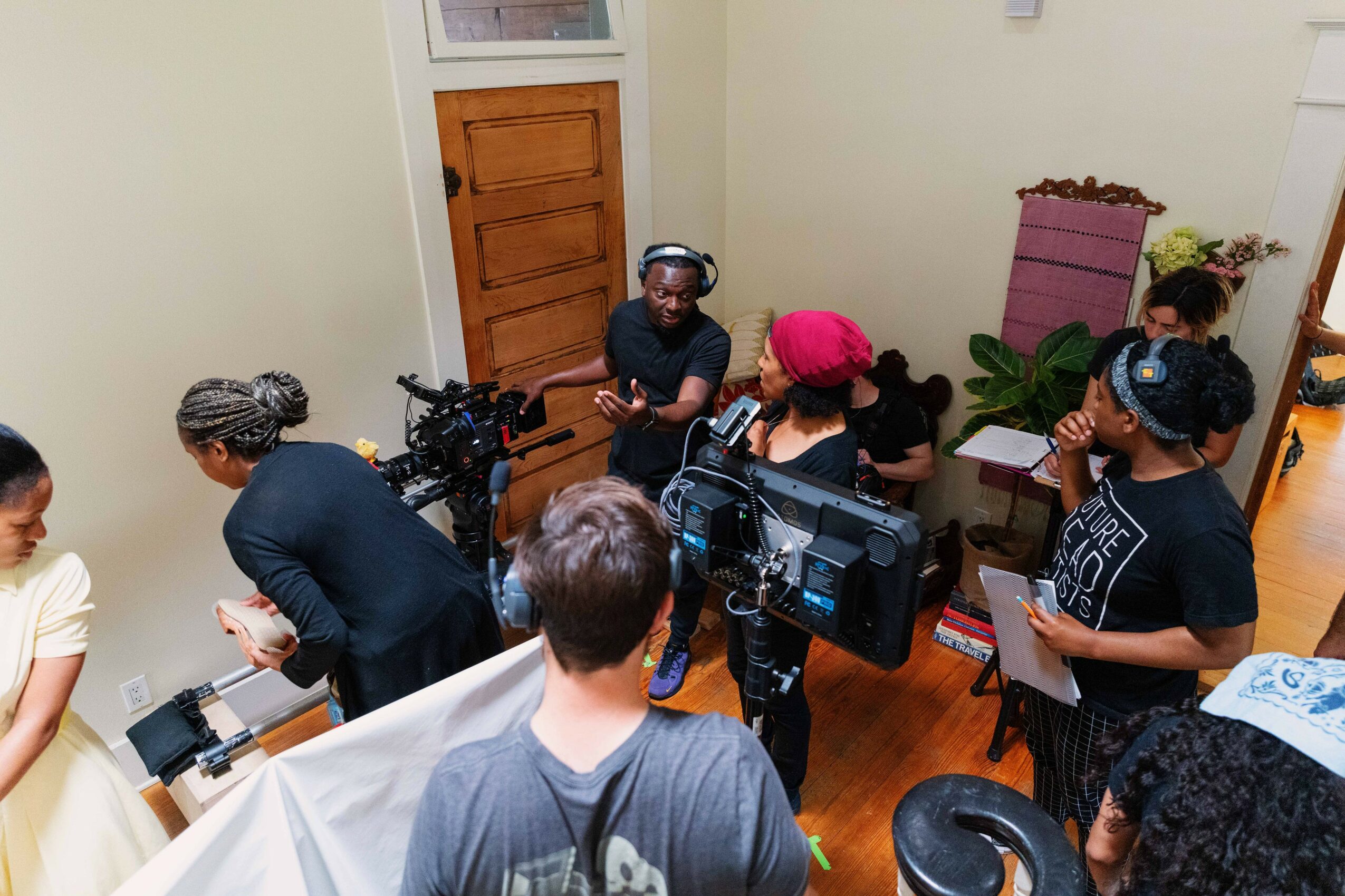 People gather in a room with a door, some using cameras and equipment, others talking or preparing items, with plants and furniture visible.