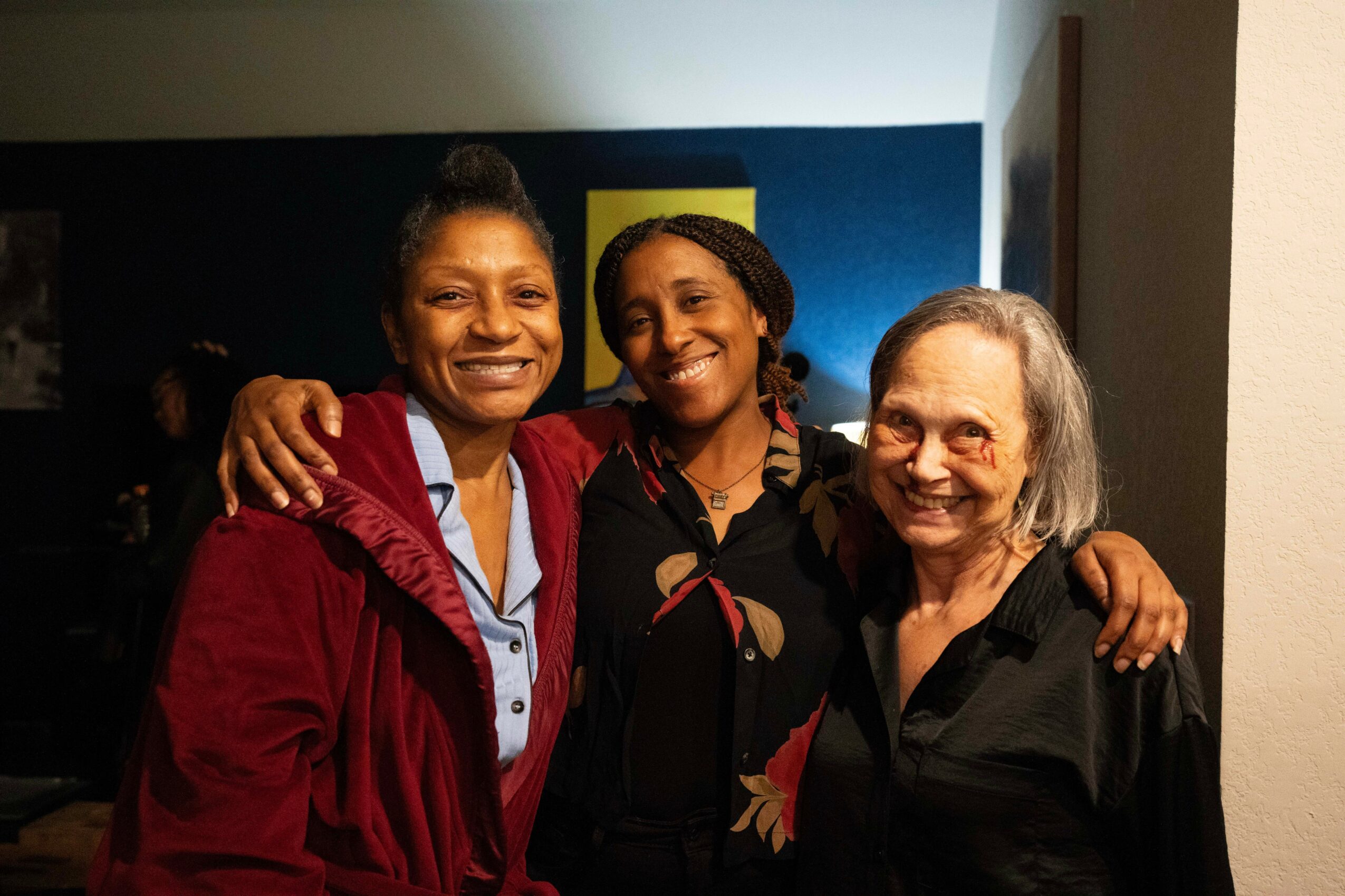 Three women smiling and hugging indoors, standing close together, with a dark background and a yellow and blue wall behind them.
