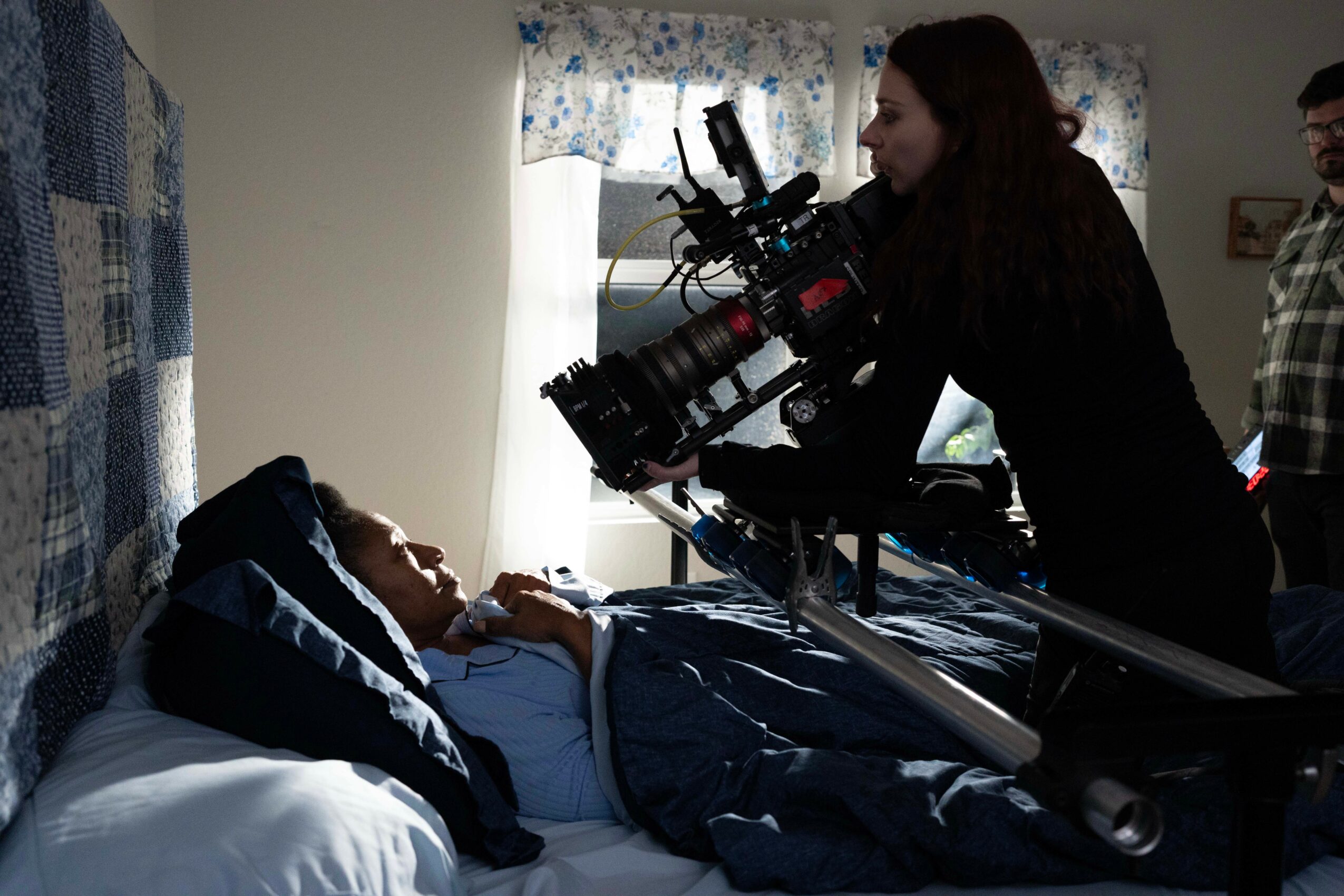 Person lying in bed being filmed by a camera operator in a room with floral curtains.