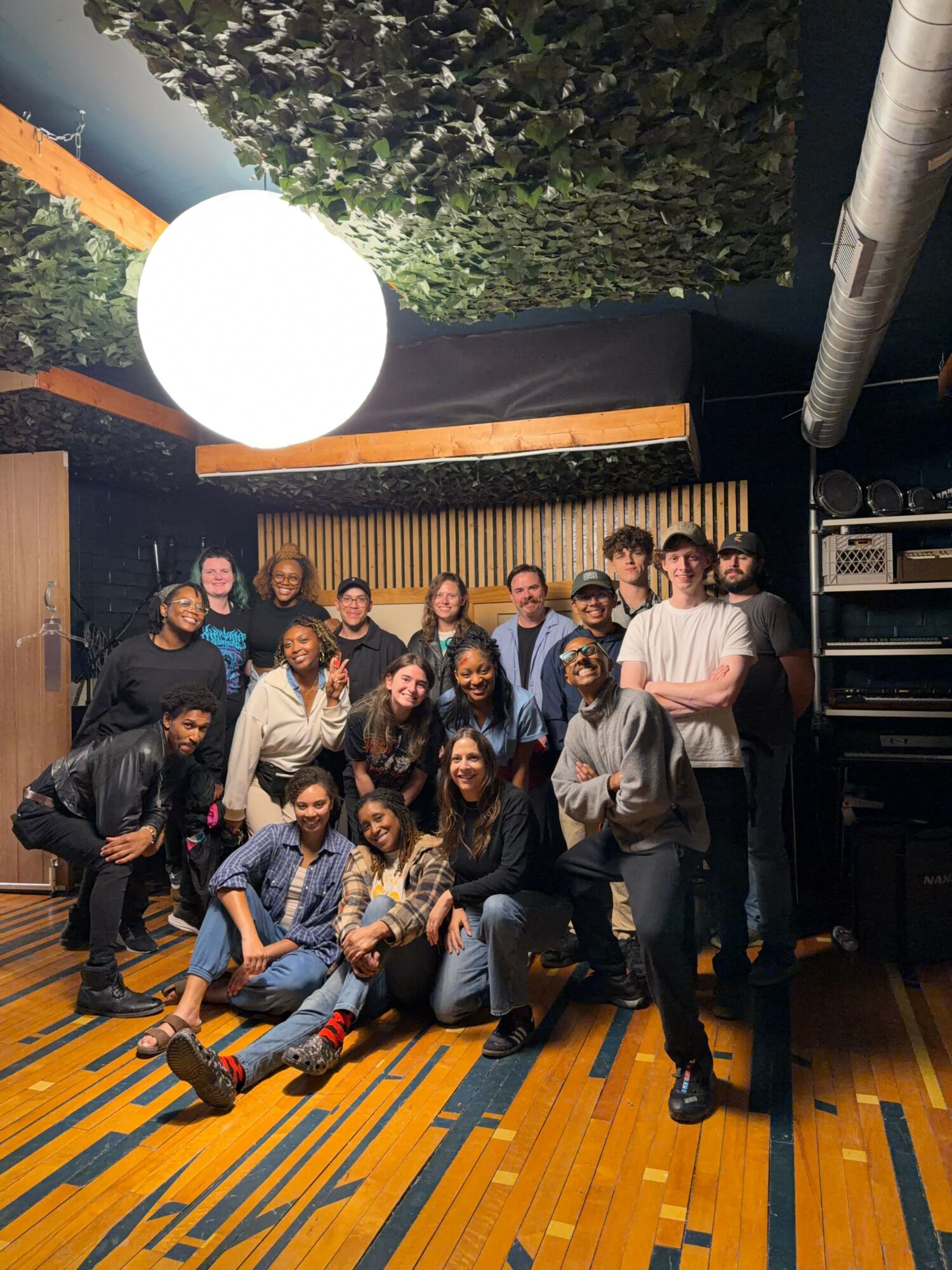 Group of people posing indoors with a black and orange background, some sitting and some standing, in a casual setting.