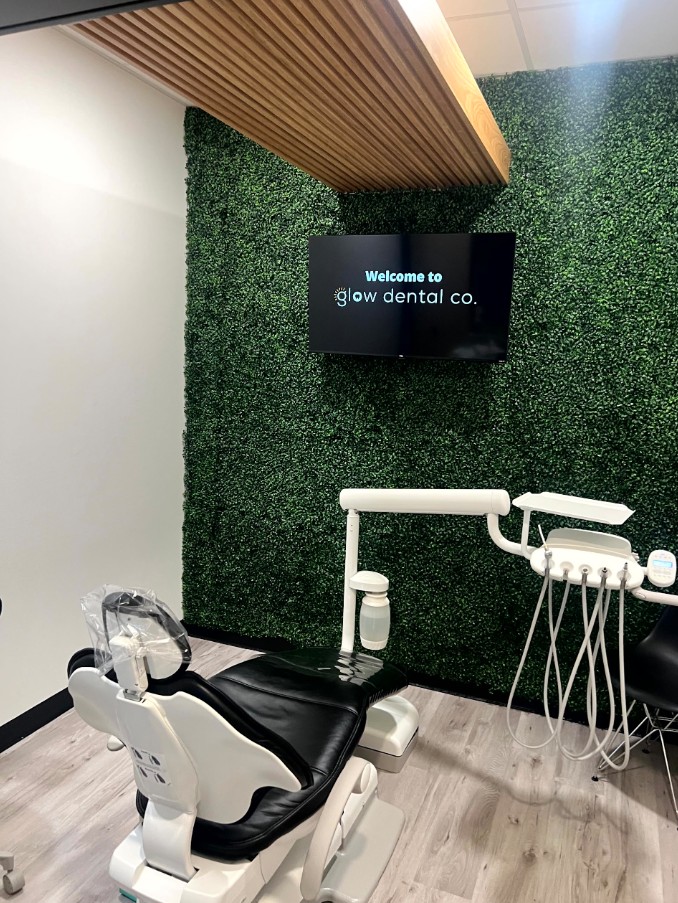 Dental clinic room with green wall, mounted screen, dental chair, and equipment, ceiling with wooden paneling.