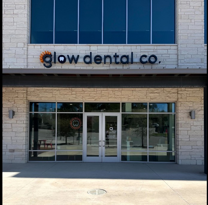 Dental office entrance with glass doors and windows, sign reads 'glow dental co.', brick exterior, reflection of street outside.