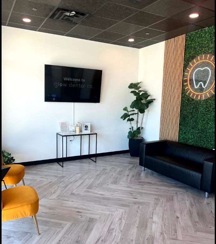 Modern dental office waiting area with a wall-mounted TV, a black sofa, a potted plant, and a decorative wall with a tooth logo.