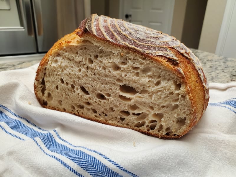 A slice of rustic bread with a thick crust and airy interior on a white cloth with blue stripes.