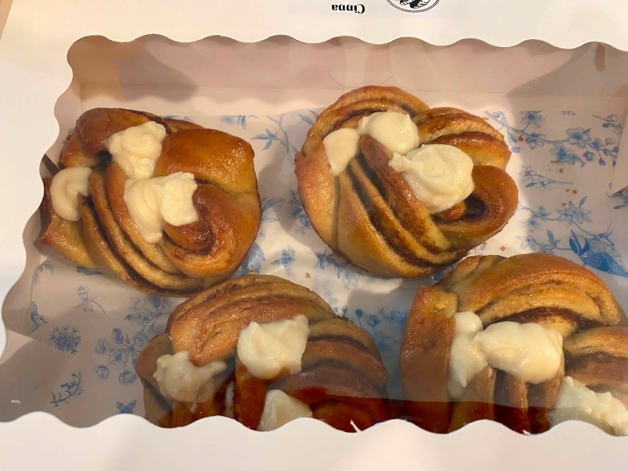 Four cinnamon rolls with icing in a box, on a blue floral paper lining.