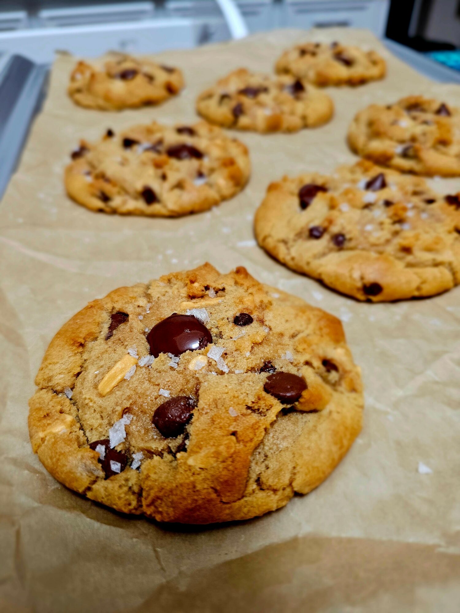 Six chocolate chip cookies on parchment paper, with one in the foreground and five in the background.