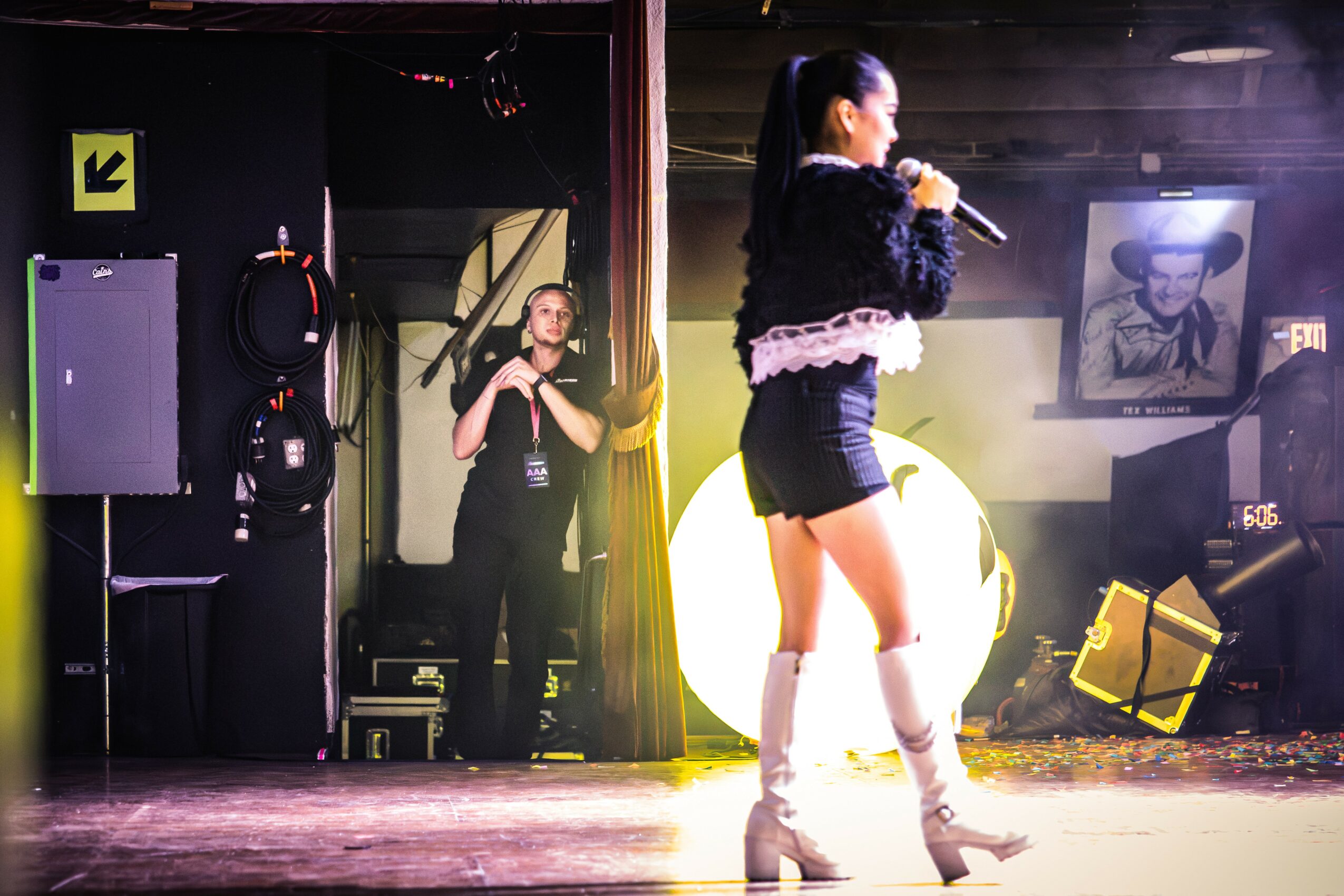 A woman performs on stage with a microphone, wearing a black outfit and white knee-high boots, in a dimly lit venue.