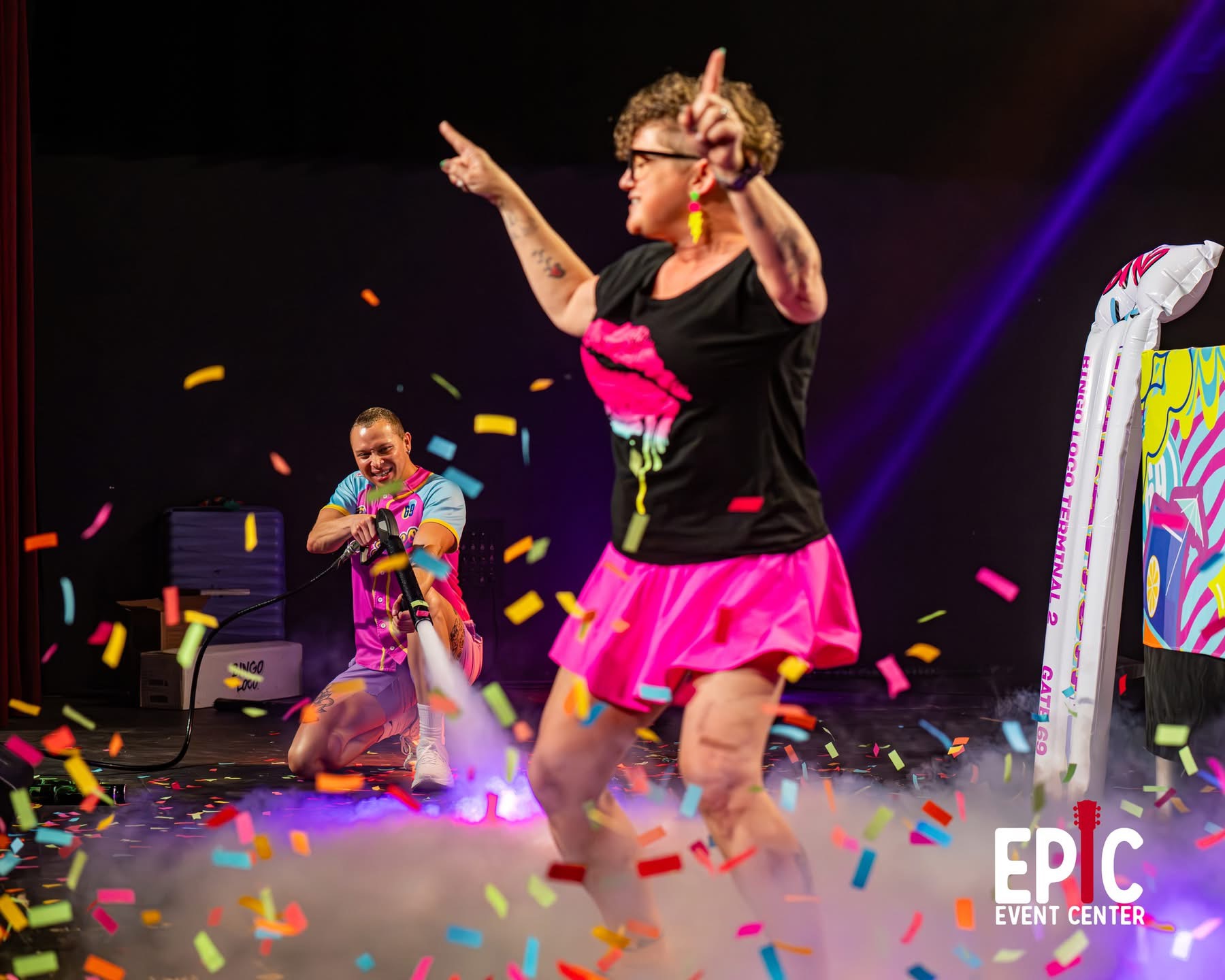 Two people dancing on stage with colorful confetti, one in a pink skirt and black top, the other in colorful clothing, at EPC Event Center.