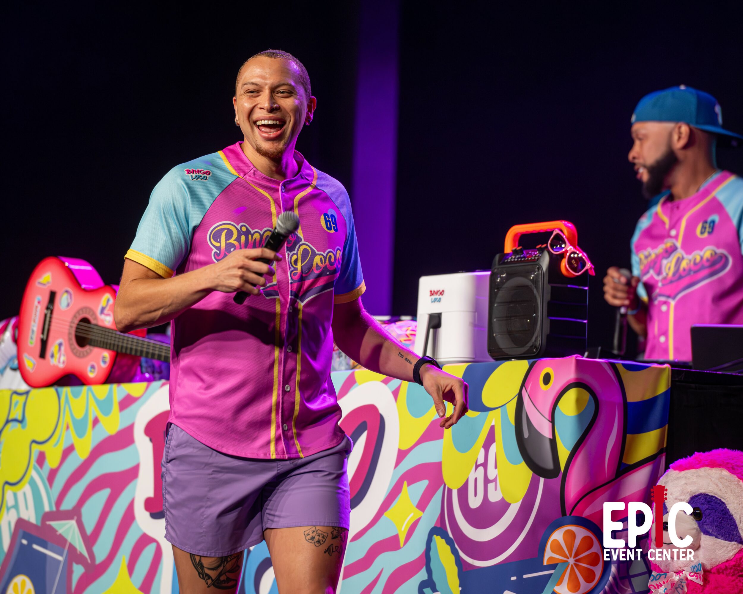 A woman in a colorful sports jersey and shorts laughs holding a microphone, with a DJ and colorful decorations in the background.